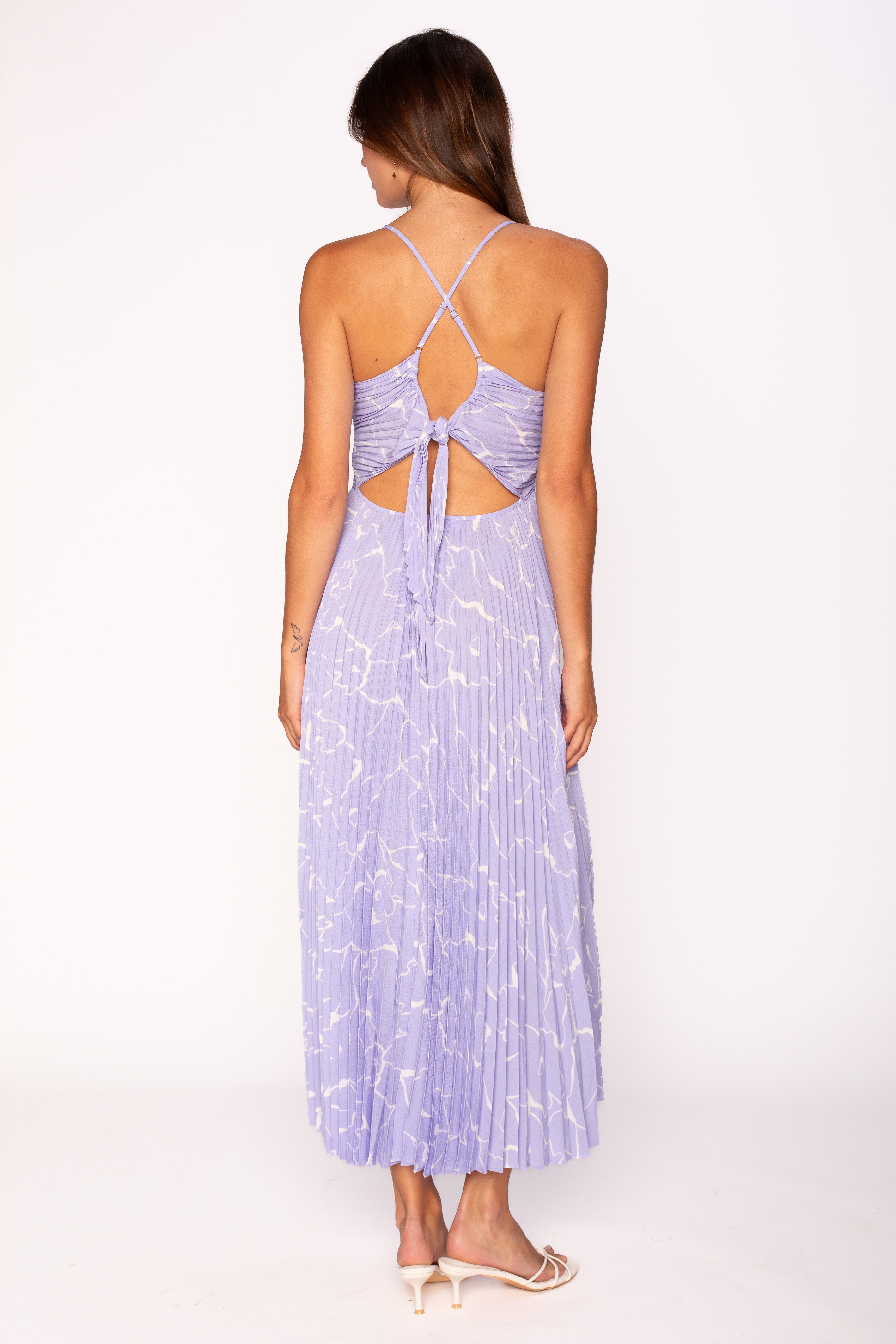 A woman faces away, wearing the ASTR Blythe Midi Dress in light purple with white patterns, featuring an open back, adjustable crisscross spaghetti straps, and a bow tie. She pairs it with light heeled sandals.