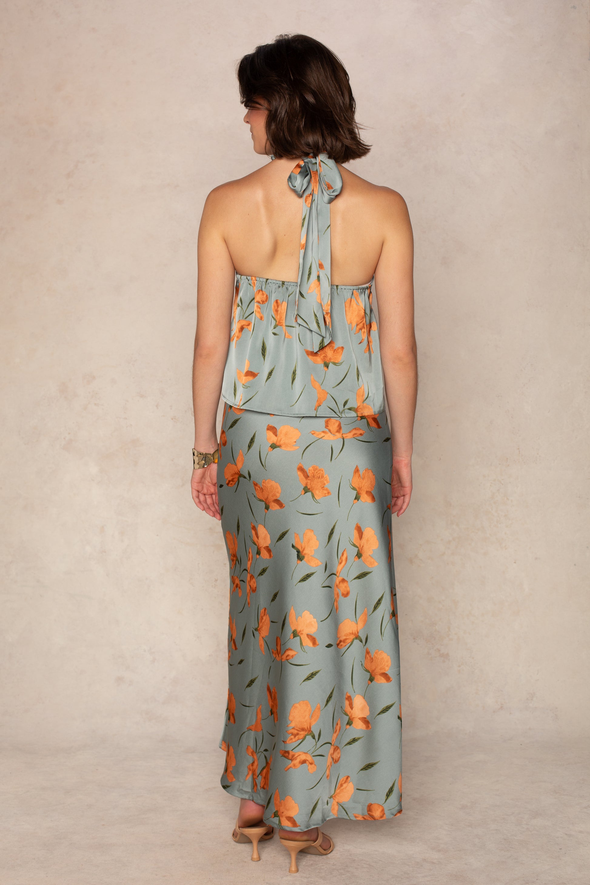 A woman stands facing away, wearing the Georgie Emilia Satin Halter—a light blue floral dress with orange prints and a high neckline. The flowy ankle-length silhouette is styled with beige heeled sandals and a bracelet on her right wrist.