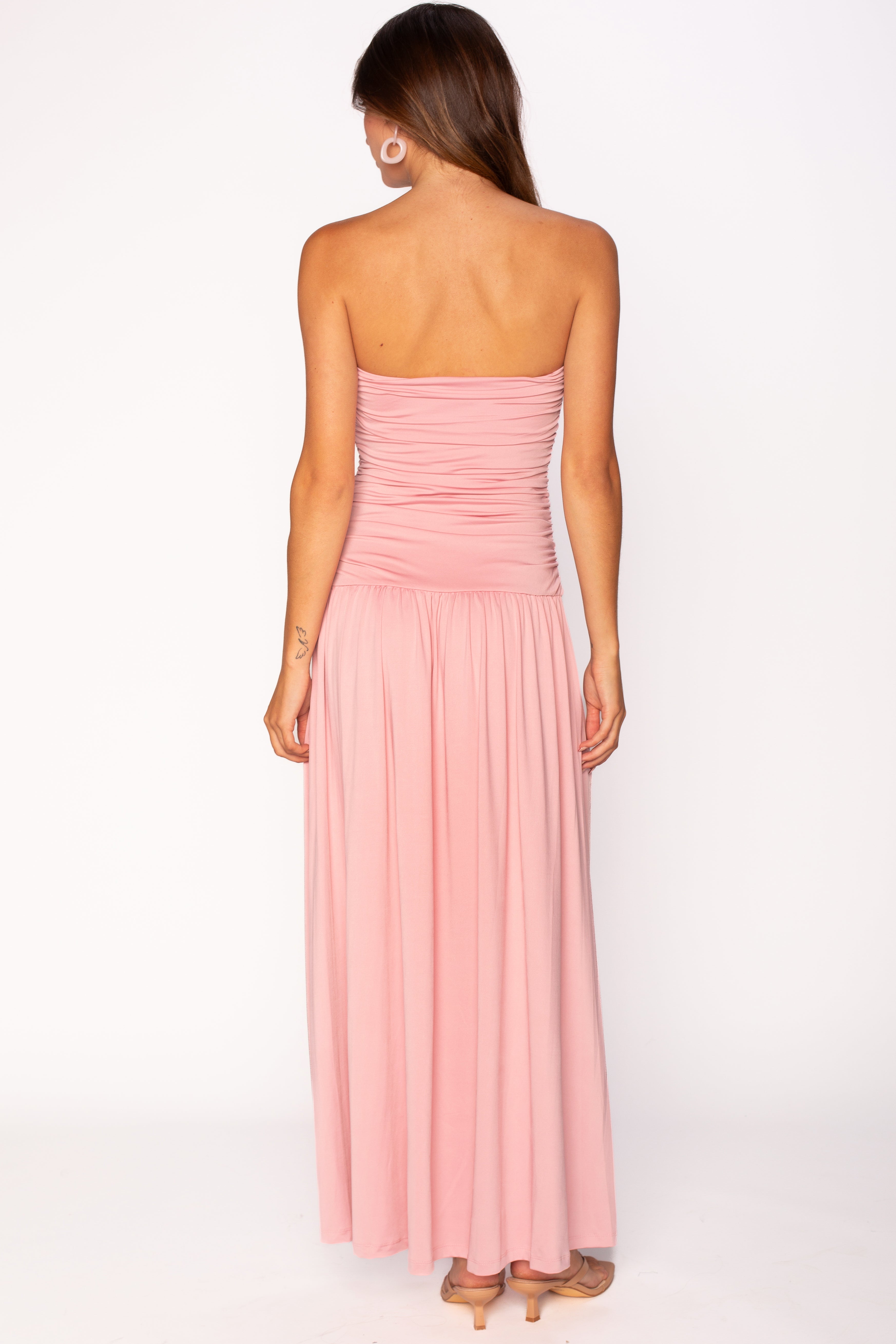 A woman with long brown hair wears the Nia Santal Midi Dress, featuring a ruched, strapless bodice and light pink skirt. She also wears hoop earrings and beige heels. The background is plain white.