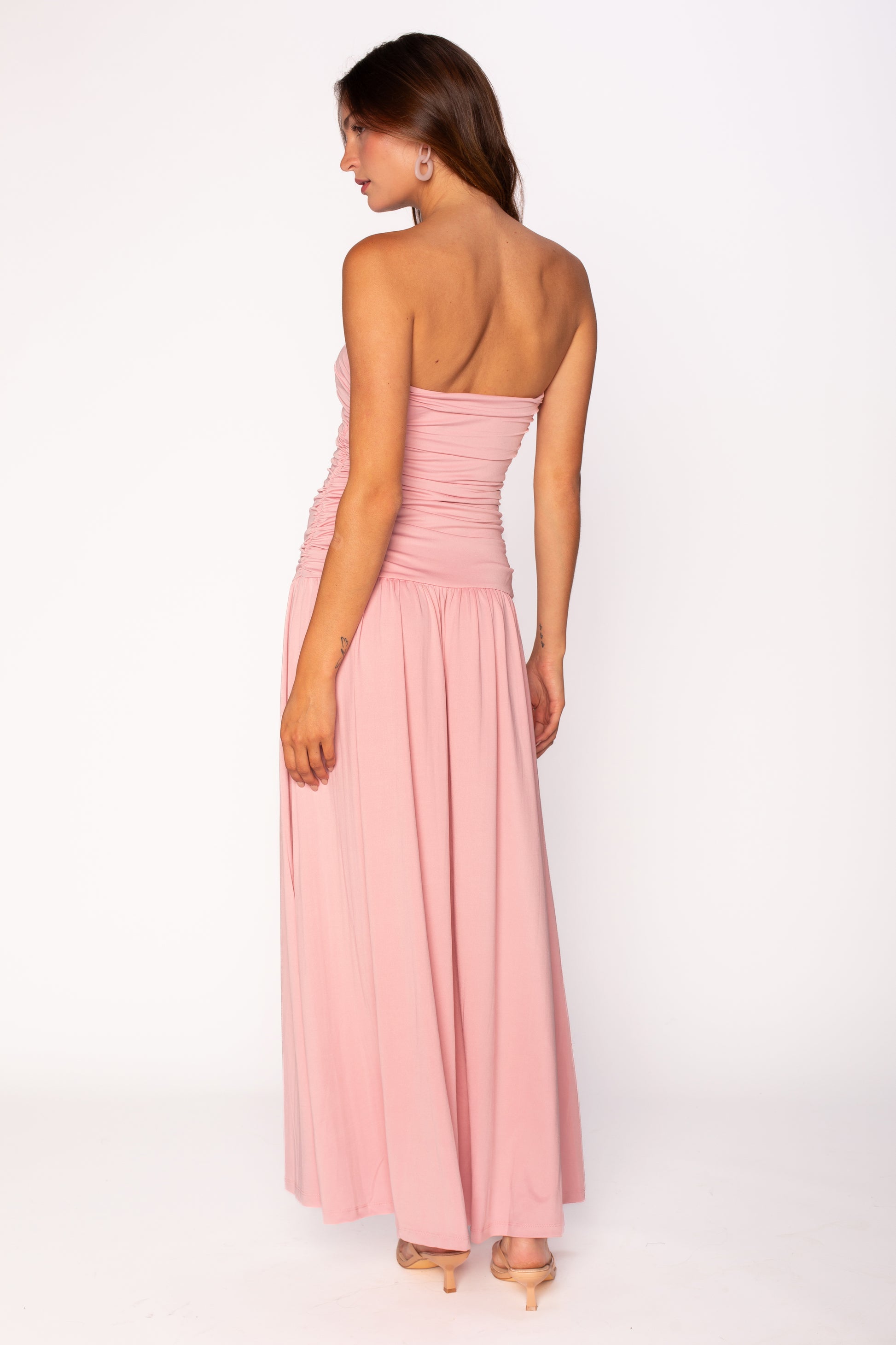 A woman in the Nia Santal Midi Dress, featuring a strapless ruched bodice, pairs it with nude heels and stands facing away while glancing over her shoulder against a plain white background.