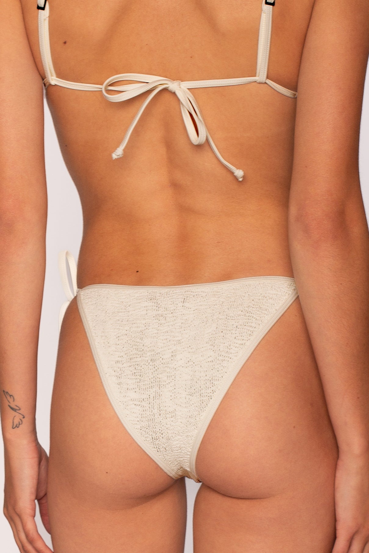 A close-up of a person’s lower back and hips shows them wearing the Bond-Eye Chalk Two Tone Luana Triangle Top with matching bikini bottoms. Adjustable straps are tied at the back, and a small butterfly tattoo is visible on their left arm.