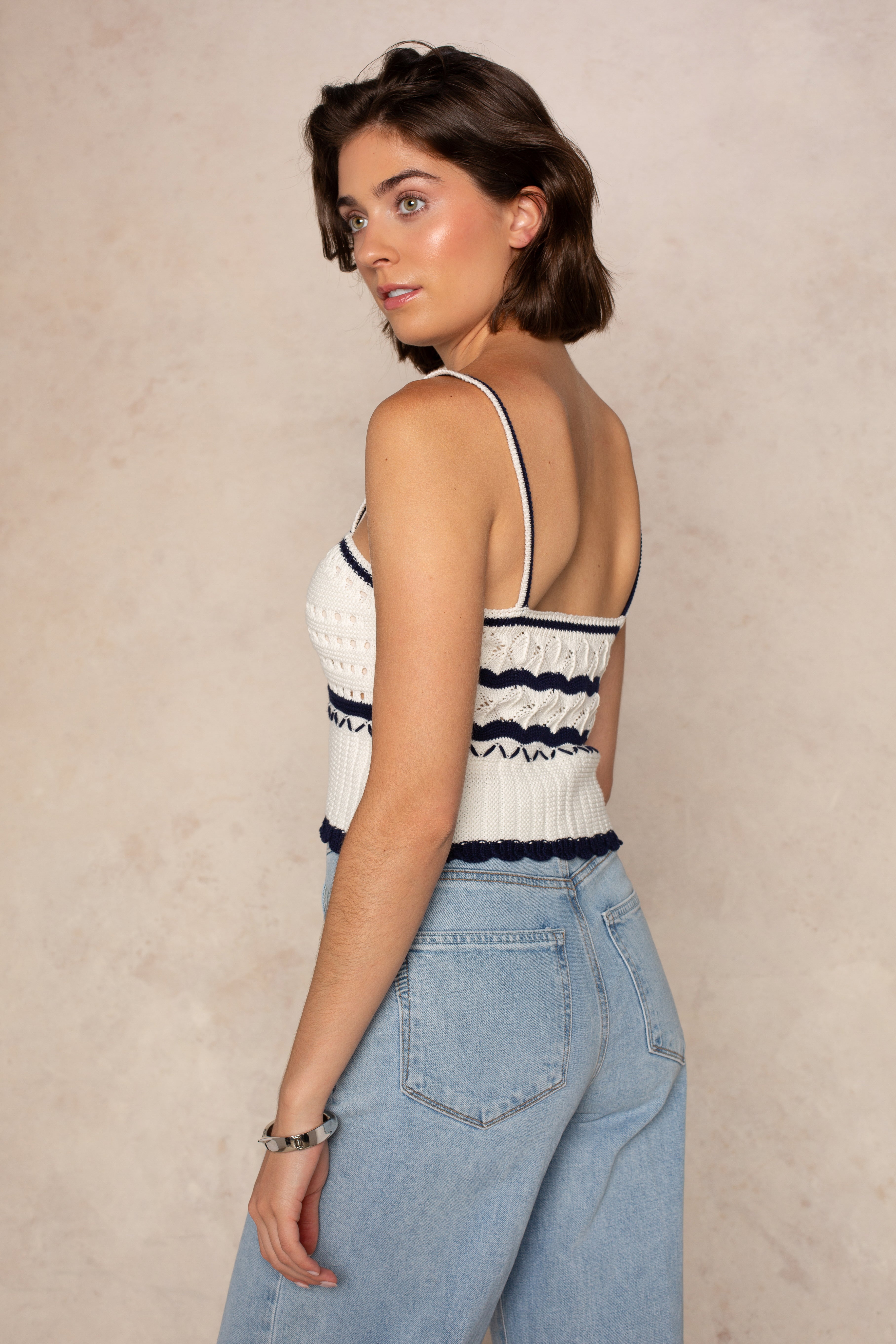 A woman with short brown hair models the Isla Layla Crochet Scallop Hem Tank and high-waisted light blue jeans, posing sideways against a beige textured background.