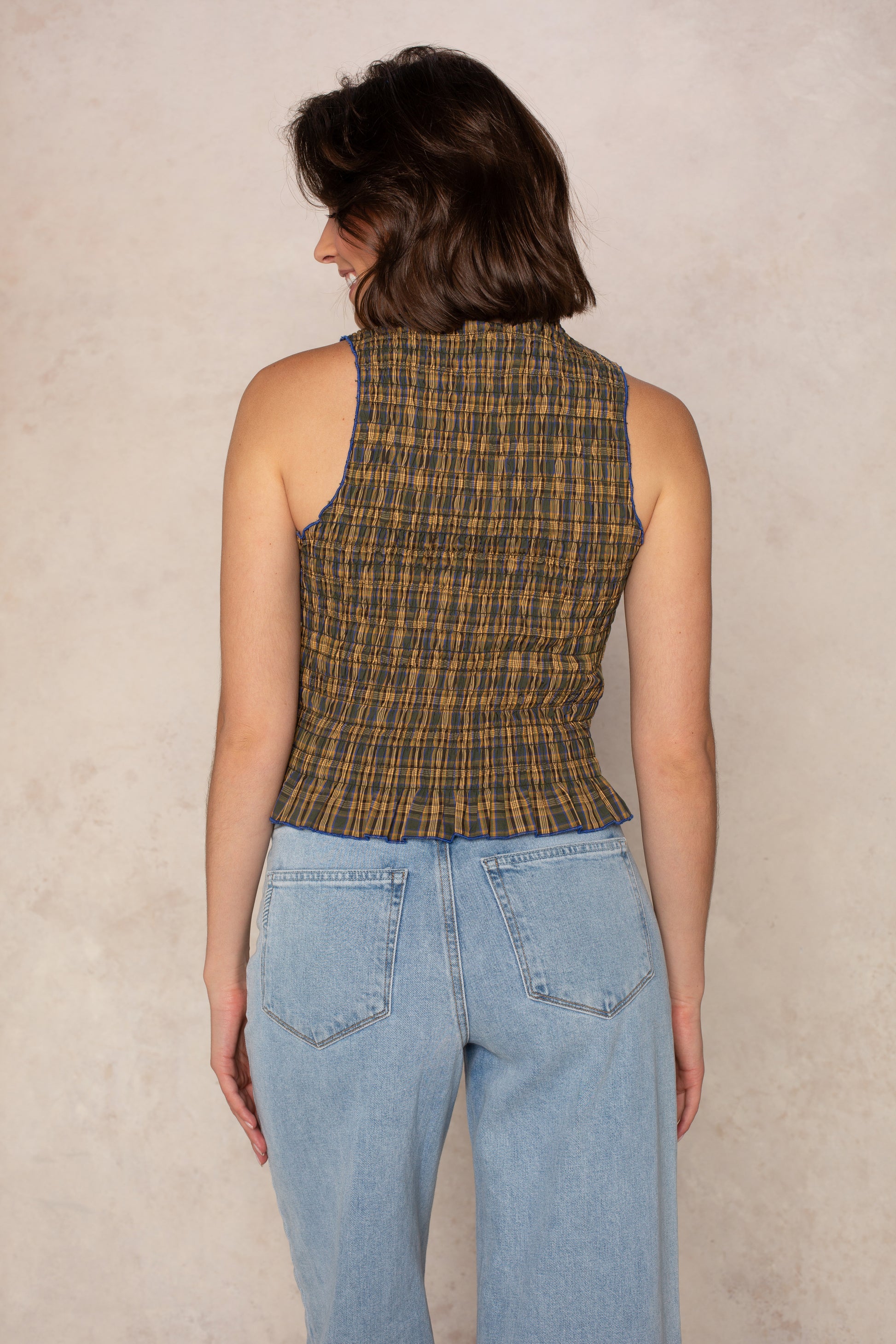 A woman with shoulder-length brown hair, seen from behind, wears the Isla Sammie Plaid Top—a sleeveless smocked-bodice plaid piece—with light blue jeans, posed against a neutral textured background.