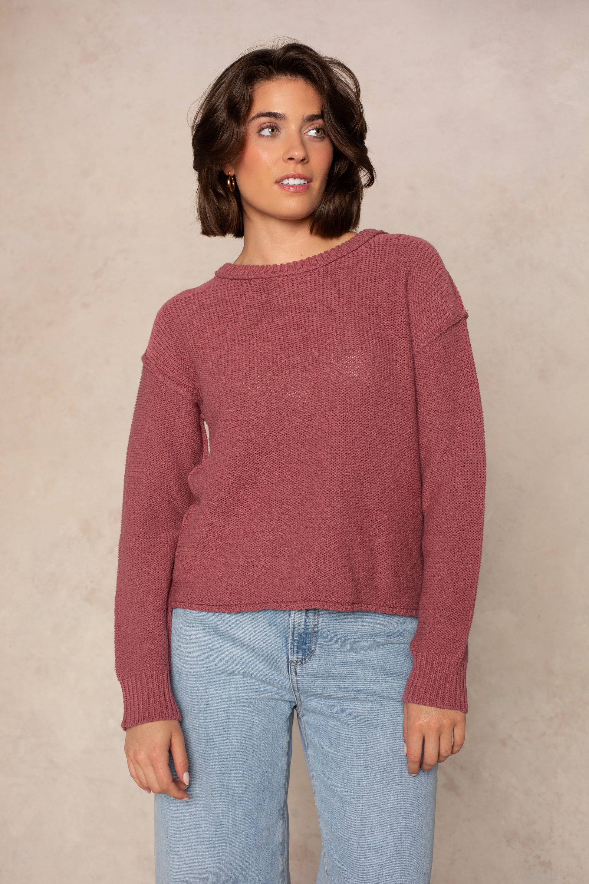 A person wearing the Isla Emmie Sweater in mauve, featuring a soft knit fabric and crew neckline, paired with light blue jeans, stands against a beige textured background and gazes slightly to the side with a relaxed expression.