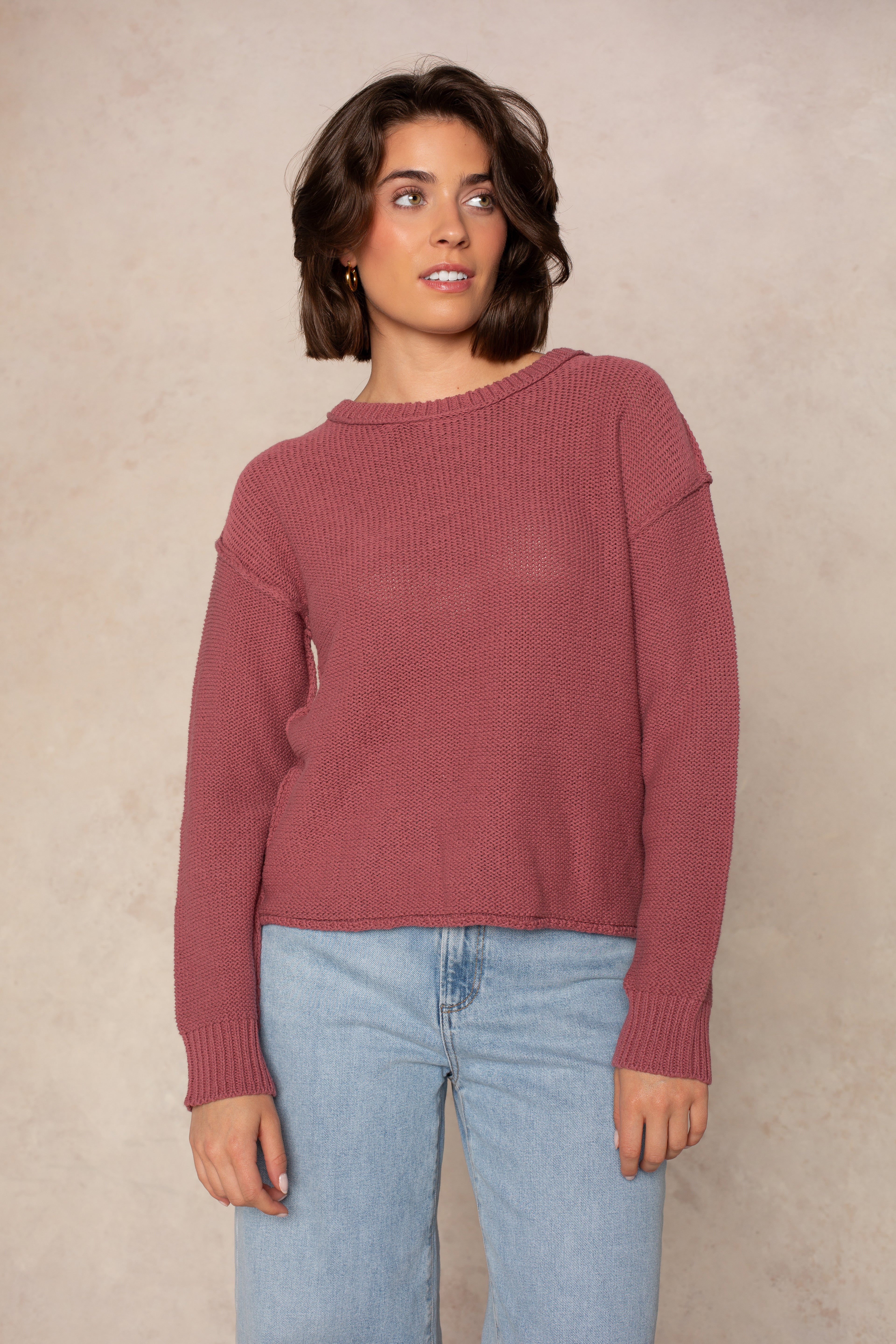 A person wearing the Isla Emmie Sweater in mauve, featuring a soft knit fabric and crew neckline, paired with light blue jeans, stands against a beige textured background and gazes slightly to the side with a relaxed expression.