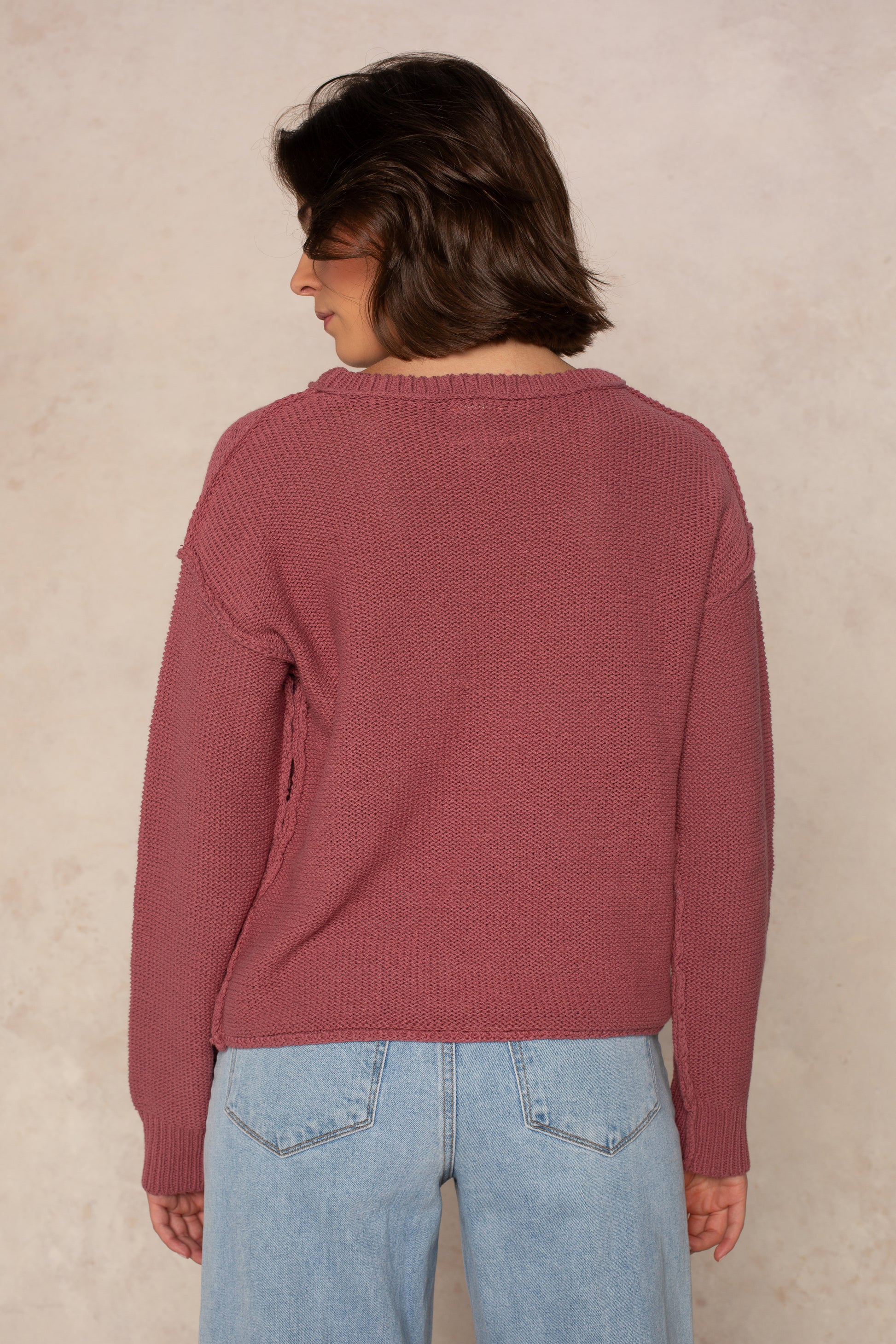 A person with short brown hair is seen from behind, wearing the Isla Emmie Sweater in mauve—a relaxed fit, crew-neck style—paired with light blue jeans and standing against a light beige textured background.