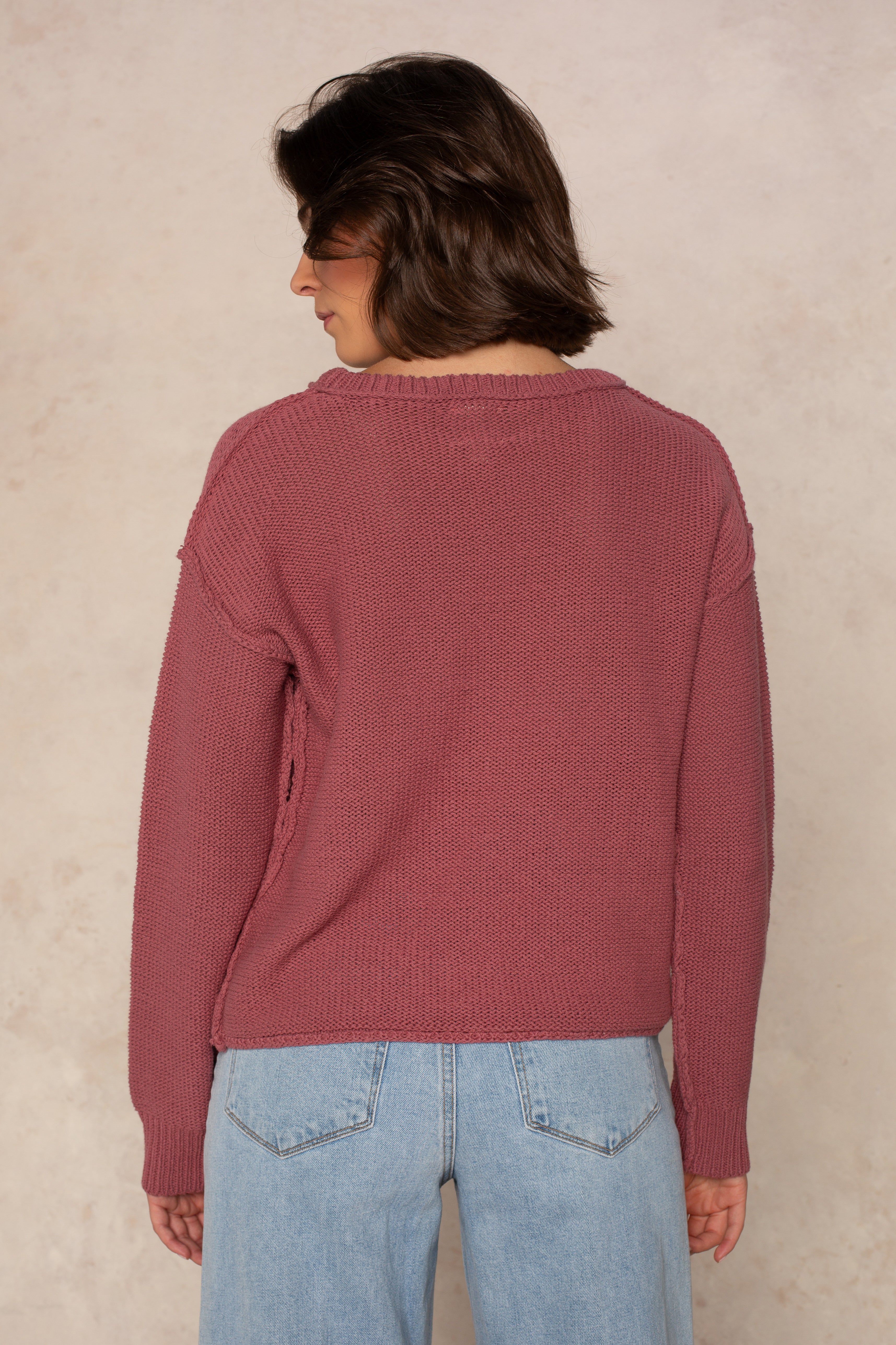 A person with short brown hair is seen from behind, wearing the Isla Emmie Sweater in mauve—a relaxed fit, crew-neck style—paired with light blue jeans and standing against a light beige textured background.