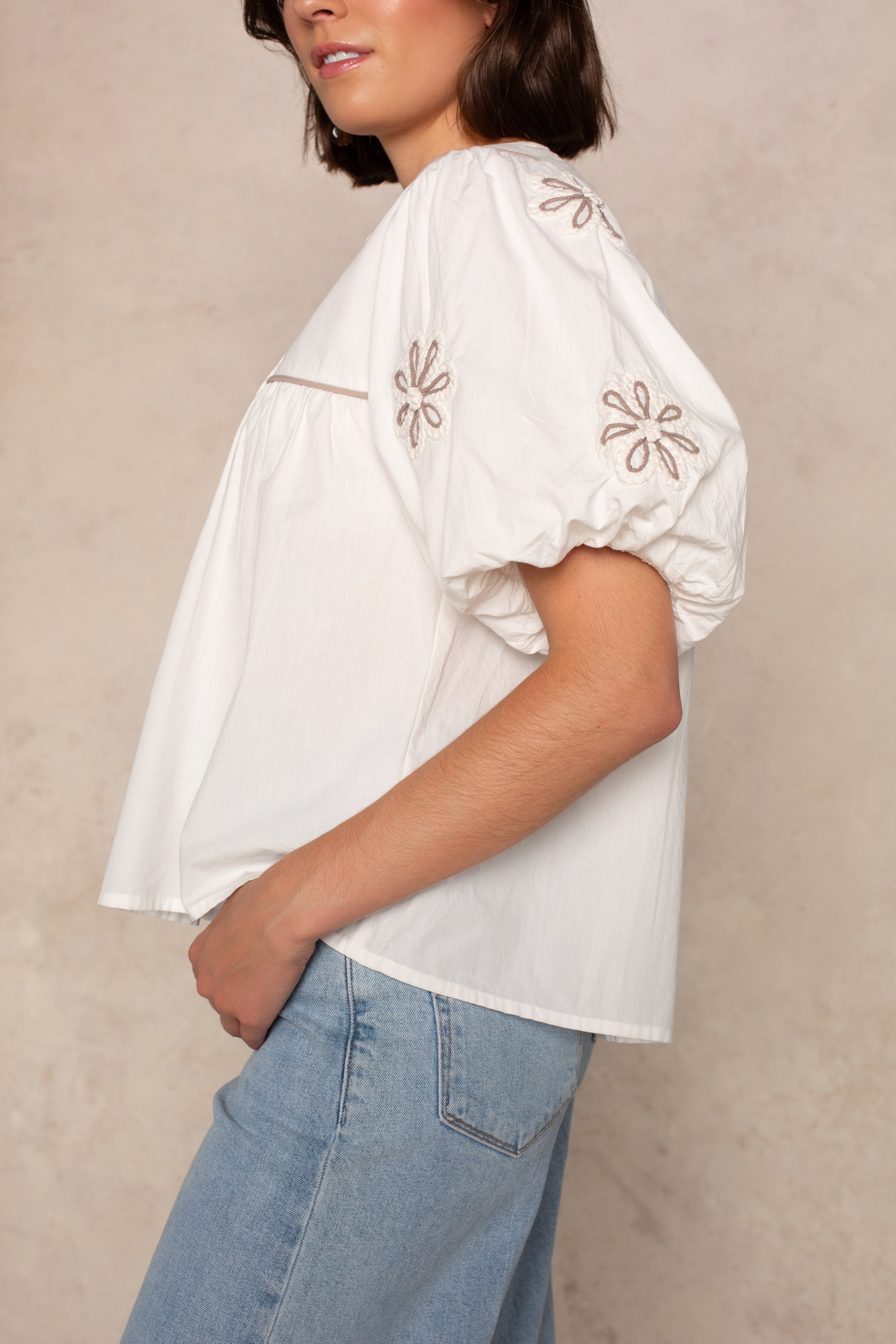 A woman models the THML Short Sleeve Tassle Top—featuring puff sleeves and floral embroidery—with light blue jeans, standing sideways against a neutral backdrop.