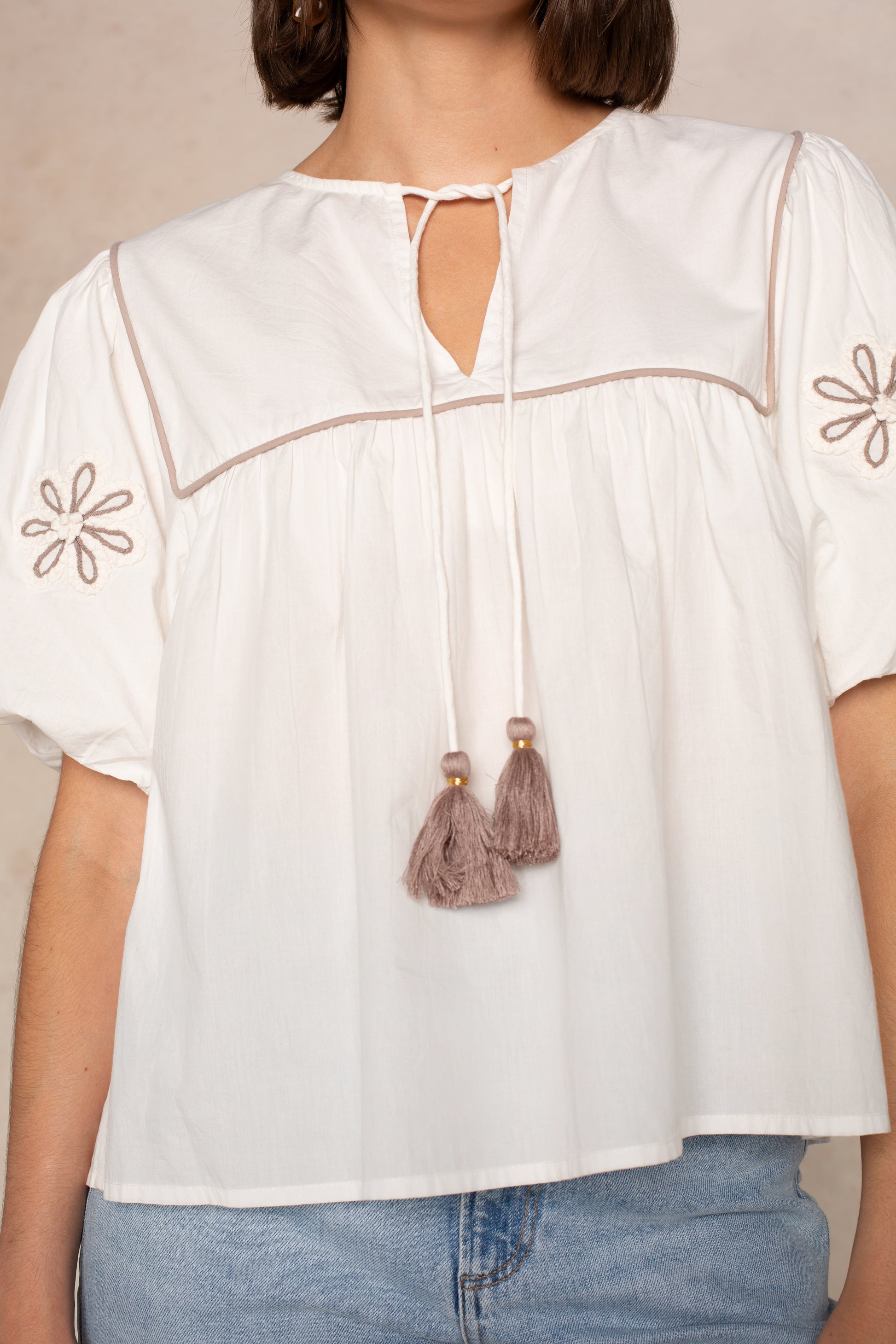 A woman wears the THML Short Sleeve Tassle Top, featuring puff sleeves, floral embroidery, a square neckline with brown piping, taupe tassel ties, paired with blue jeans.