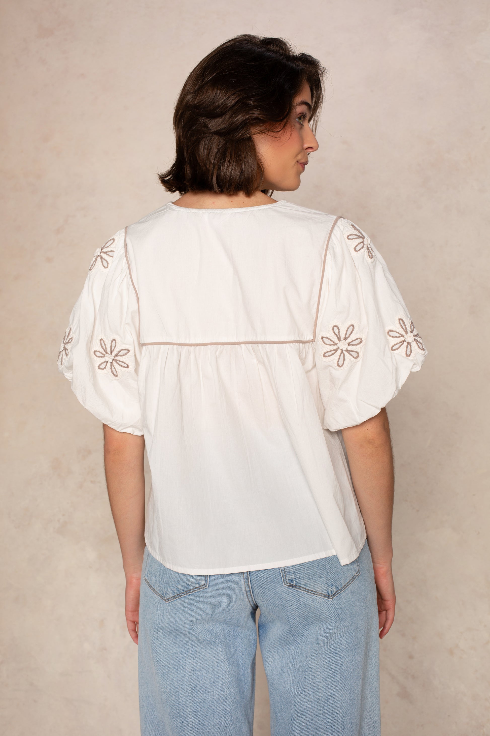 A person with short brown hair stands against a beige background, wearing the THML Short Sleeve Tassle Top—a white lightweight woven top with puff sleeves and floral embroidery—paired with light blue jeans.