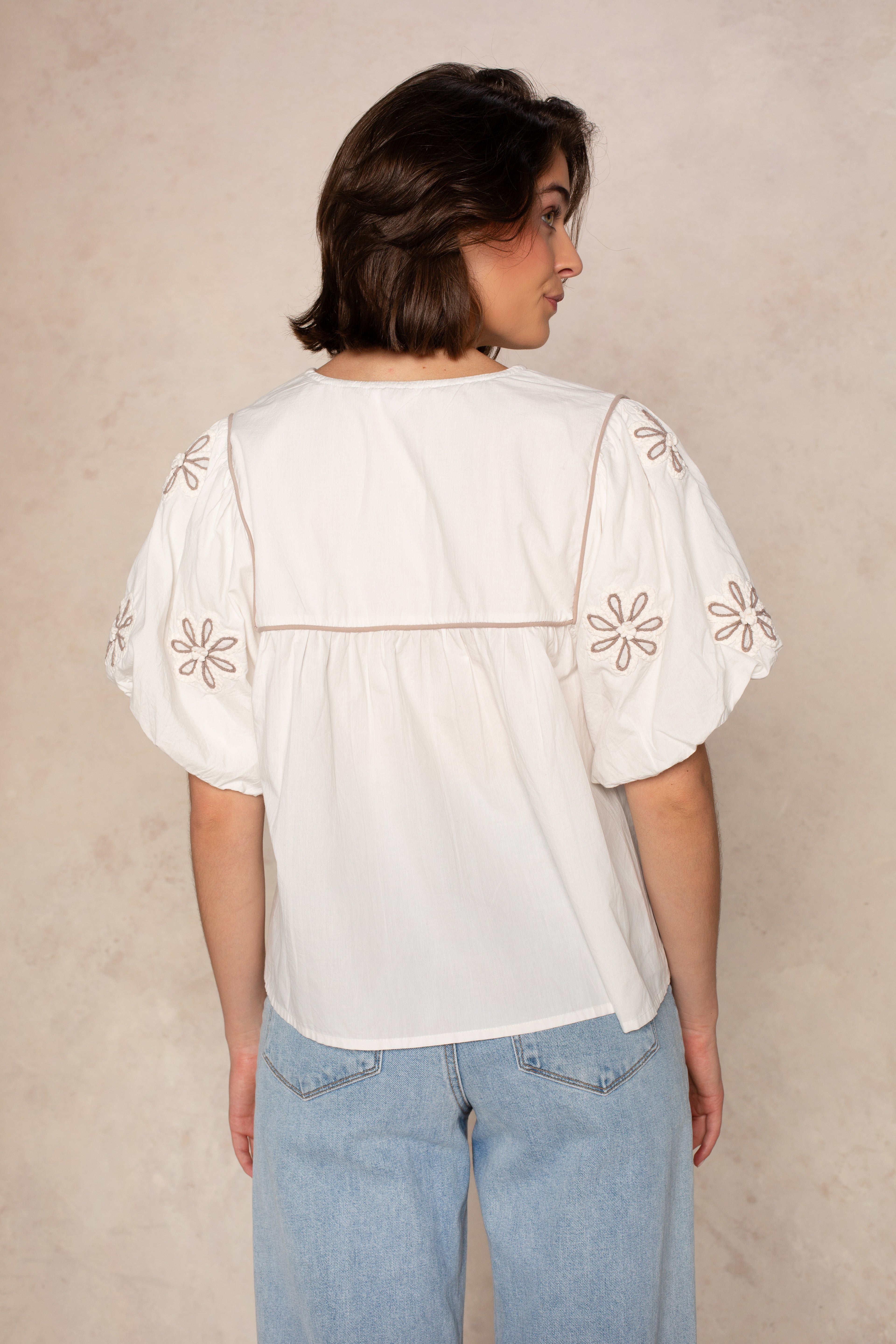 A person with short brown hair stands against a beige background, wearing the THML Short Sleeve Tassle Top—a white lightweight woven top with puff sleeves and floral embroidery—paired with light blue jeans.