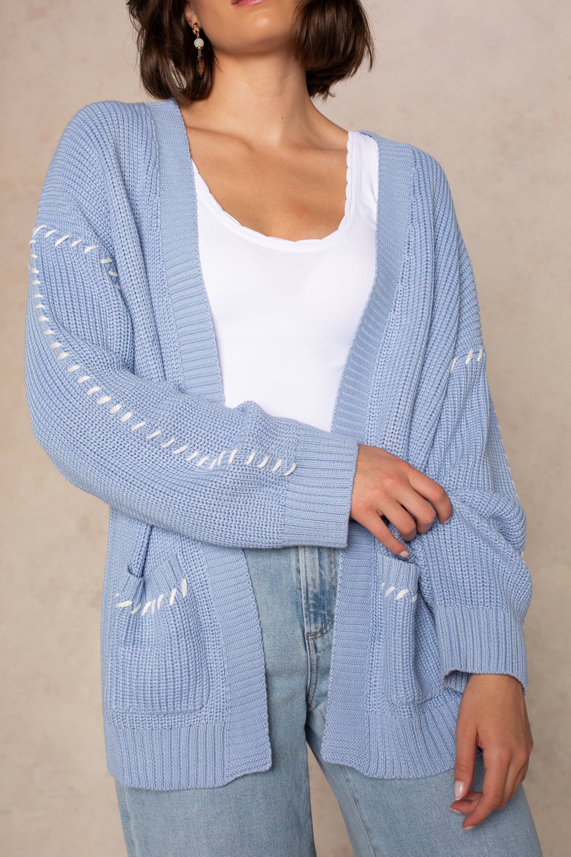 A woman stands against a beige background wearing Isla's Open Knit Oversize Cardigan in DUSTY BLUE, styled over a white top and light blue jeans. The chunky cardigan features an oversized fit, white thread detailing, two front pockets, and an open-front design.