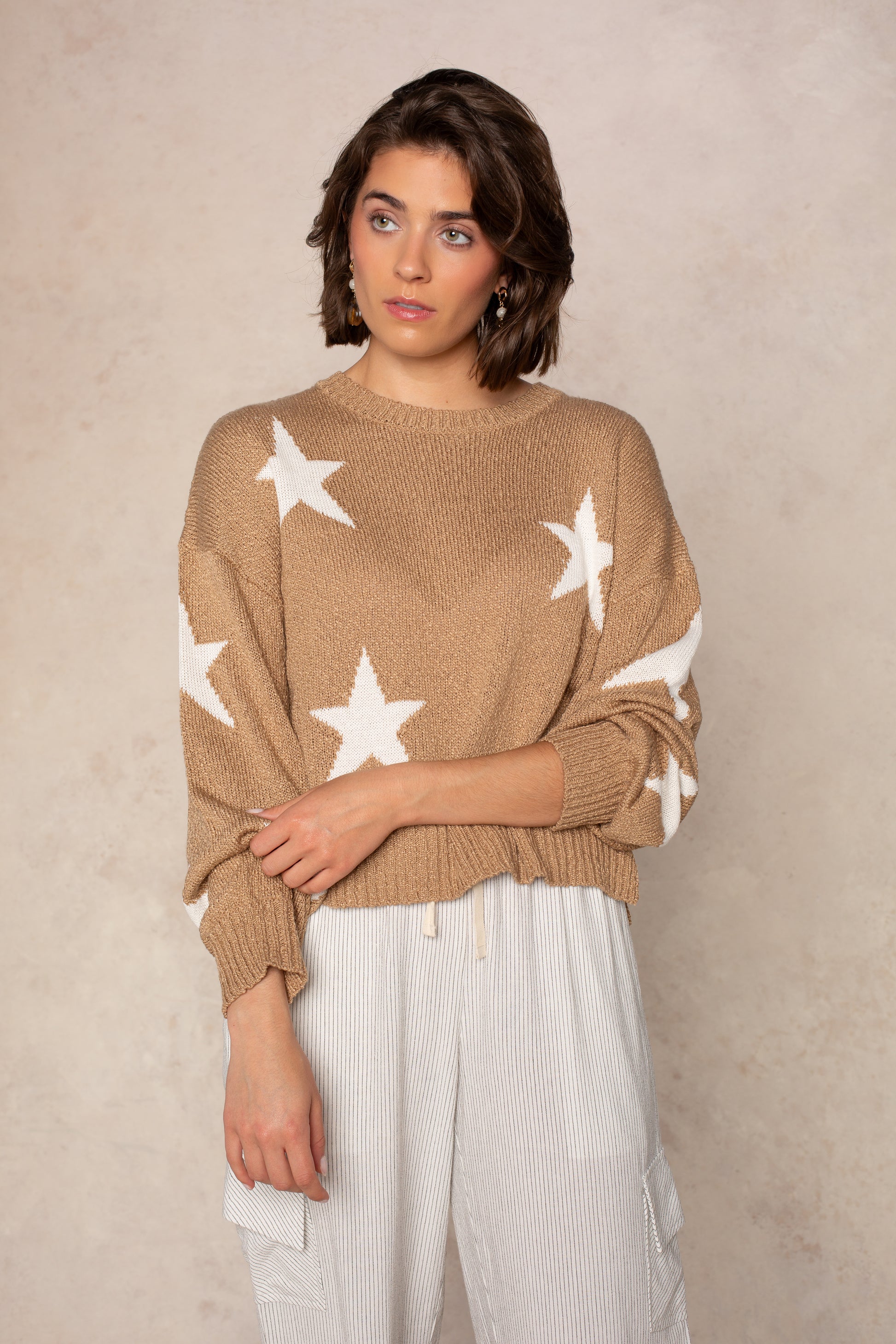 A woman with short brown hair wears Isla’s Starry Sky Sweater in BROWN and light striped pants, standing before a textured neutral background. She gazes slightly to the side with a thoughtful look.