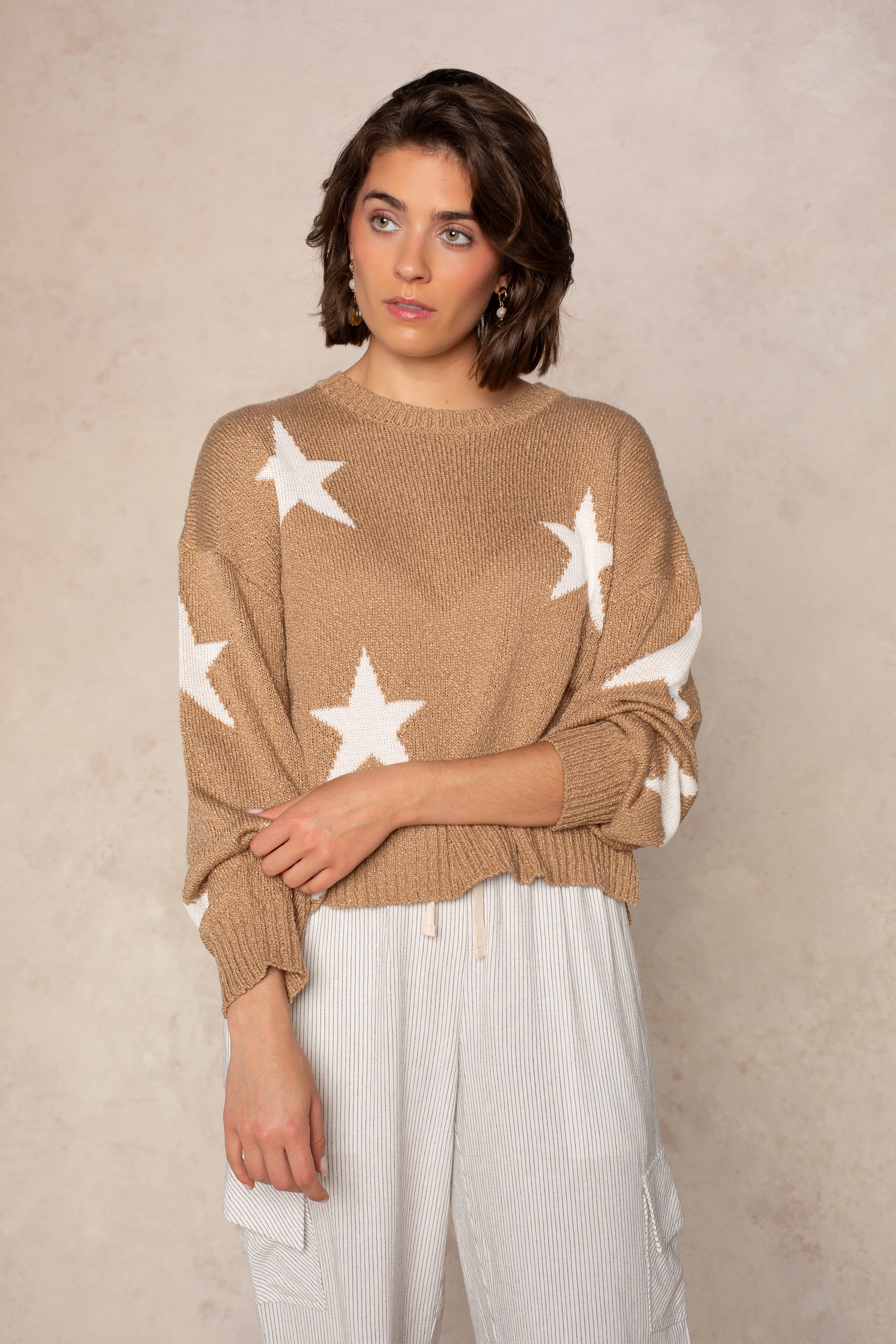 A woman with short brown hair wears Isla’s Starry Sky Sweater in BROWN and light striped pants, standing before a textured neutral background. She gazes slightly to the side with a thoughtful look.
