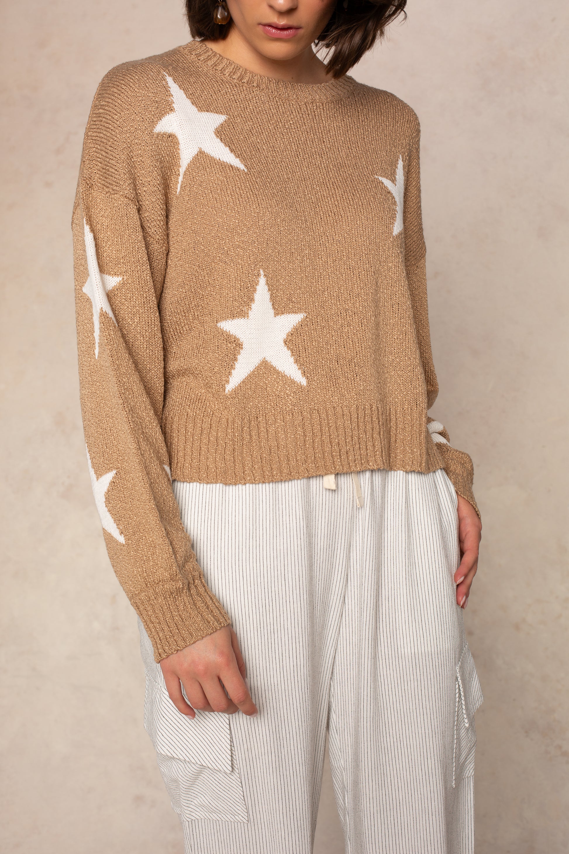 A person wears the Isla Starry Sky Sweater, a cozy knit with white stars, paired with loose light-striped pants. The soft sweater adds an everyday layer, and the face is partly out of frame against a neutral beige background.