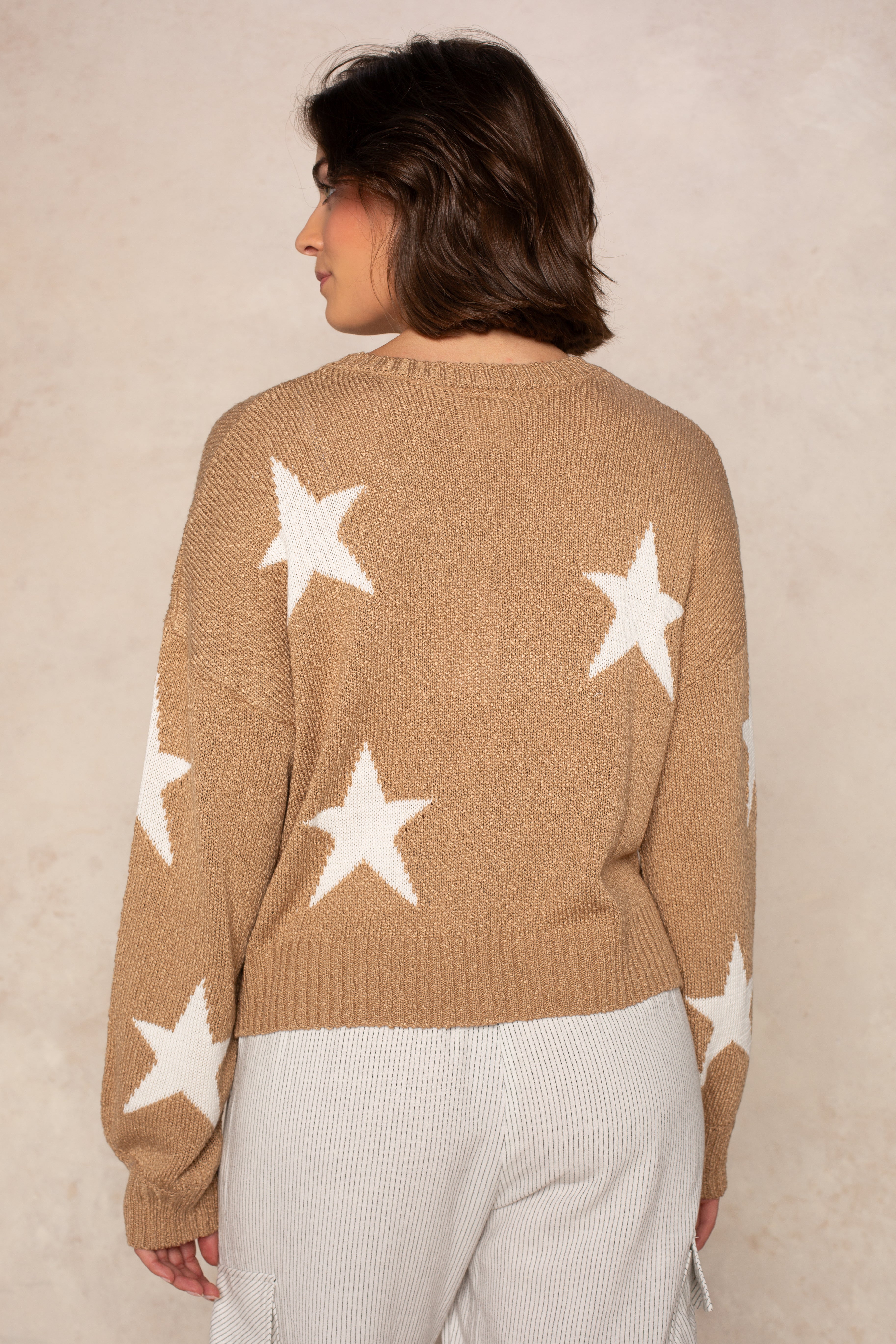 Seen from behind, a person with short brown hair wears the Isla Starry Sky Sweater—an everyday tan layer with a star pattern—paired with light striped pants, standing against a neutral background.