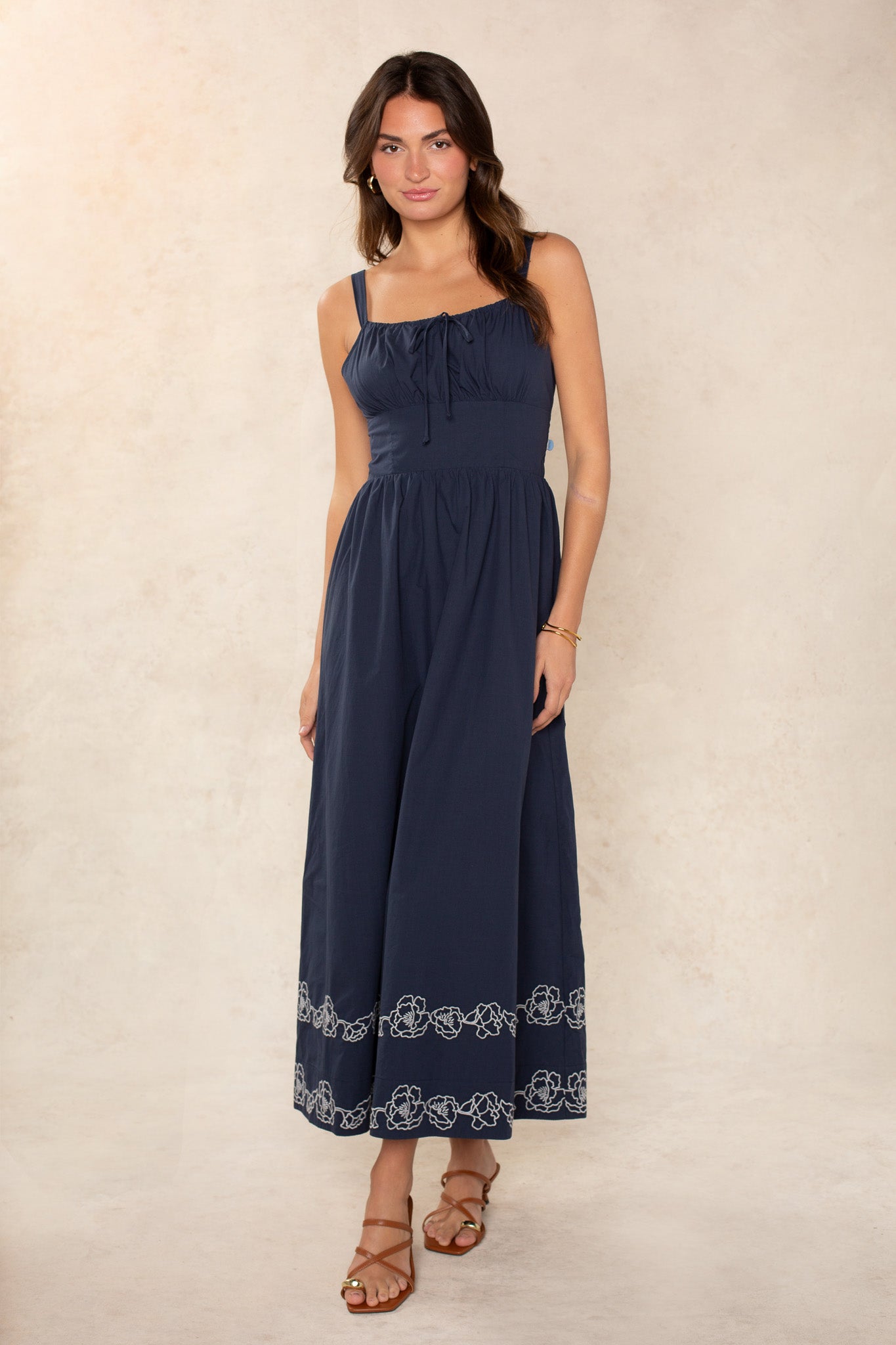 Against a light beige background, a woman wears Georgie's Hadley Embroidered Midi Dress in navy with white detailing at the hem. Her loose wavy hair, brown strappy sandals, and minimal jewelry complete her effortlessly feminine look.