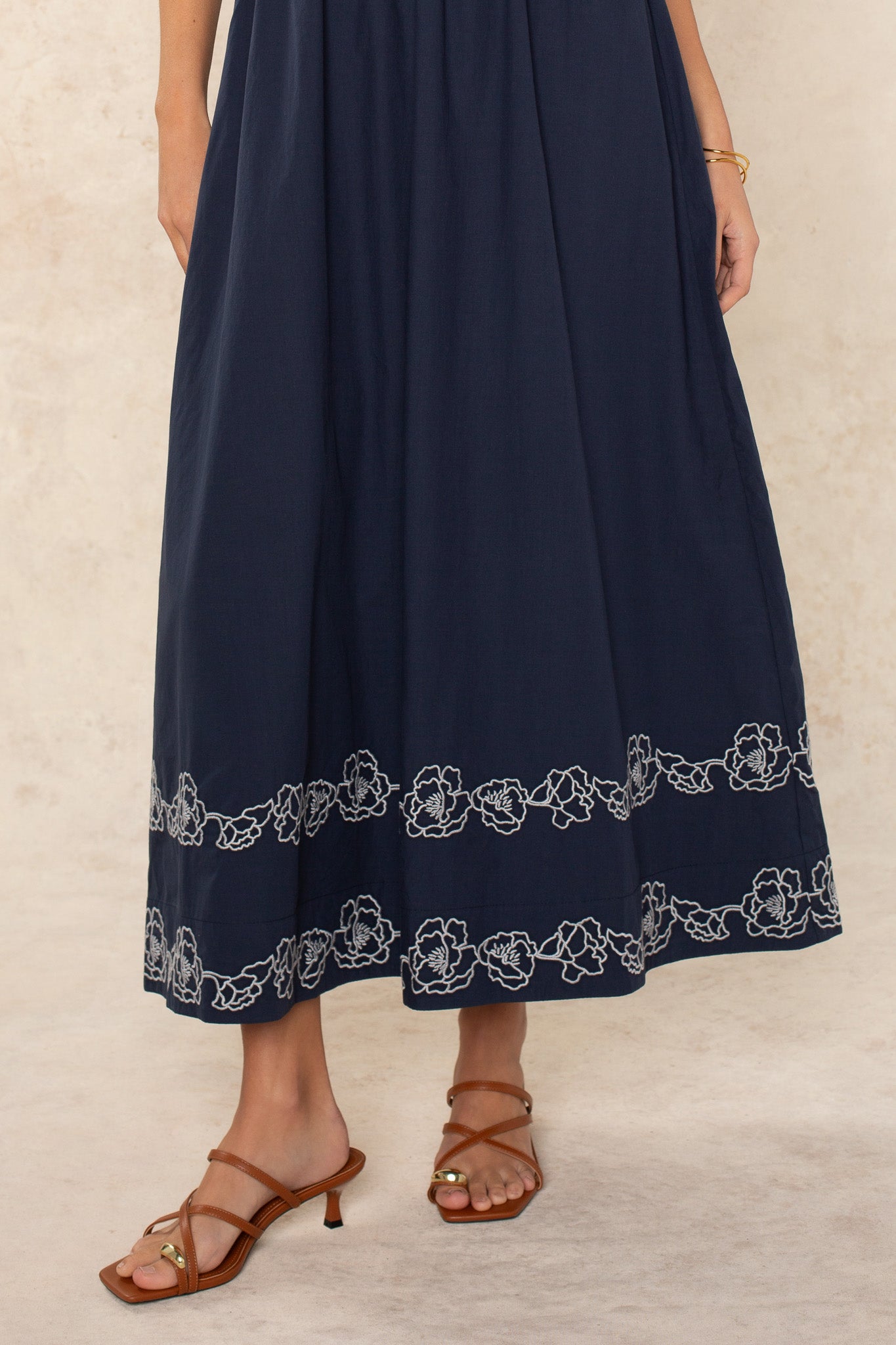 A woman models the Georgie Hadley Embroidered Midi Dress, featuring navy blue fabric with white floral embroidery at the hem. She completes the feminine look with brown strappy heeled sandals and a gold bracelet against a light background.