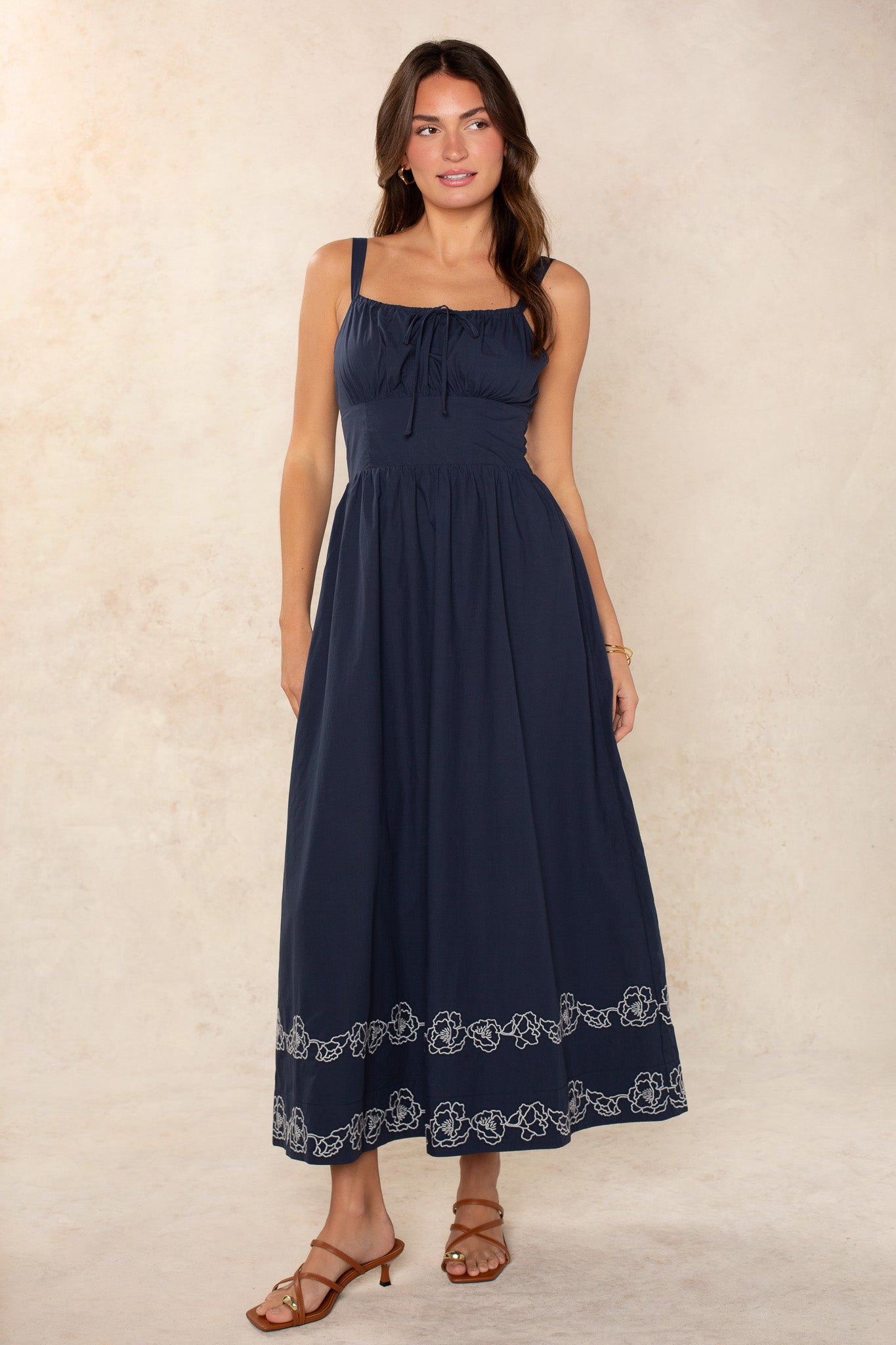 A woman with long dark hair stands against a light beige background wearing the Georgie Hadley Embroidered Midi Dress in navy, featuring white floral details at the hem. She pairs it with brown heeled sandals for a chic, feminine look.