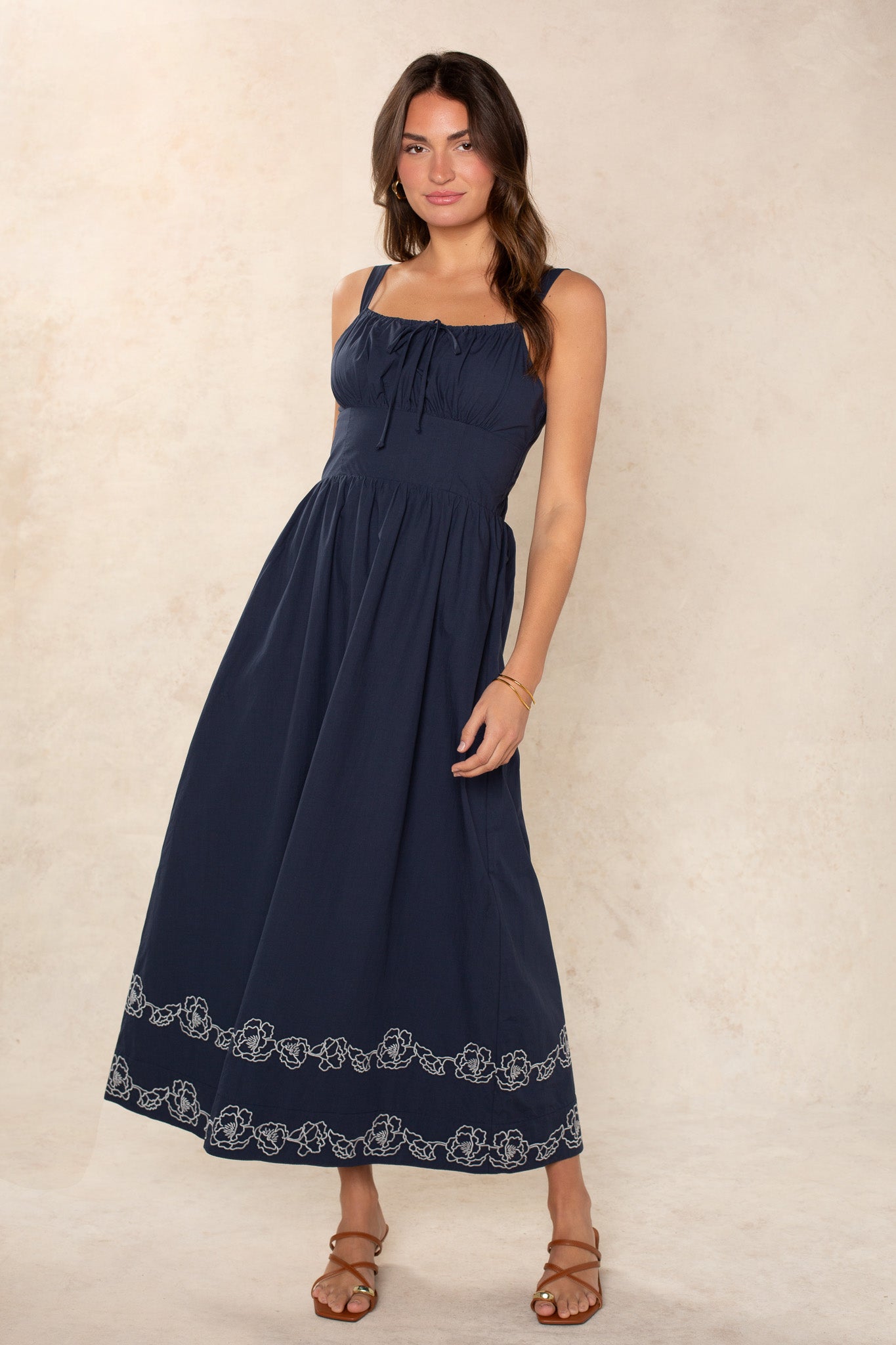 A woman poses against a light textured background in the Georgie Hadley Embroidered Midi Dress, featuring navy blue fabric with white embroidery near the hem. She completes her look with loose hair, brown sandals, and gold bracelets.