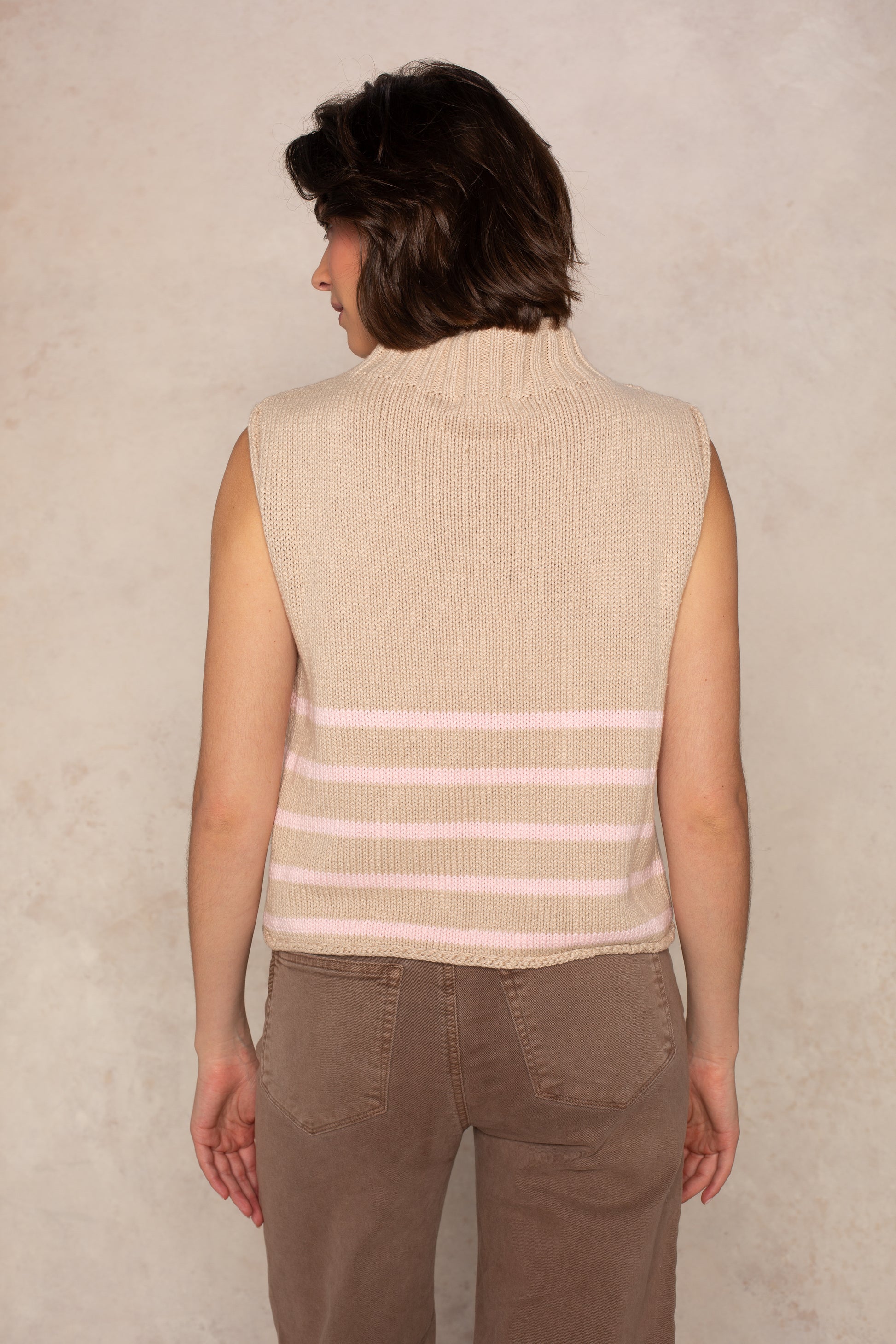 A person with short brown hair is seen from behind wearing the Georgie Grace Rolled Neck Sleeveless Sweater in Tan/Pink, paired with brown pants, against a light textured background.