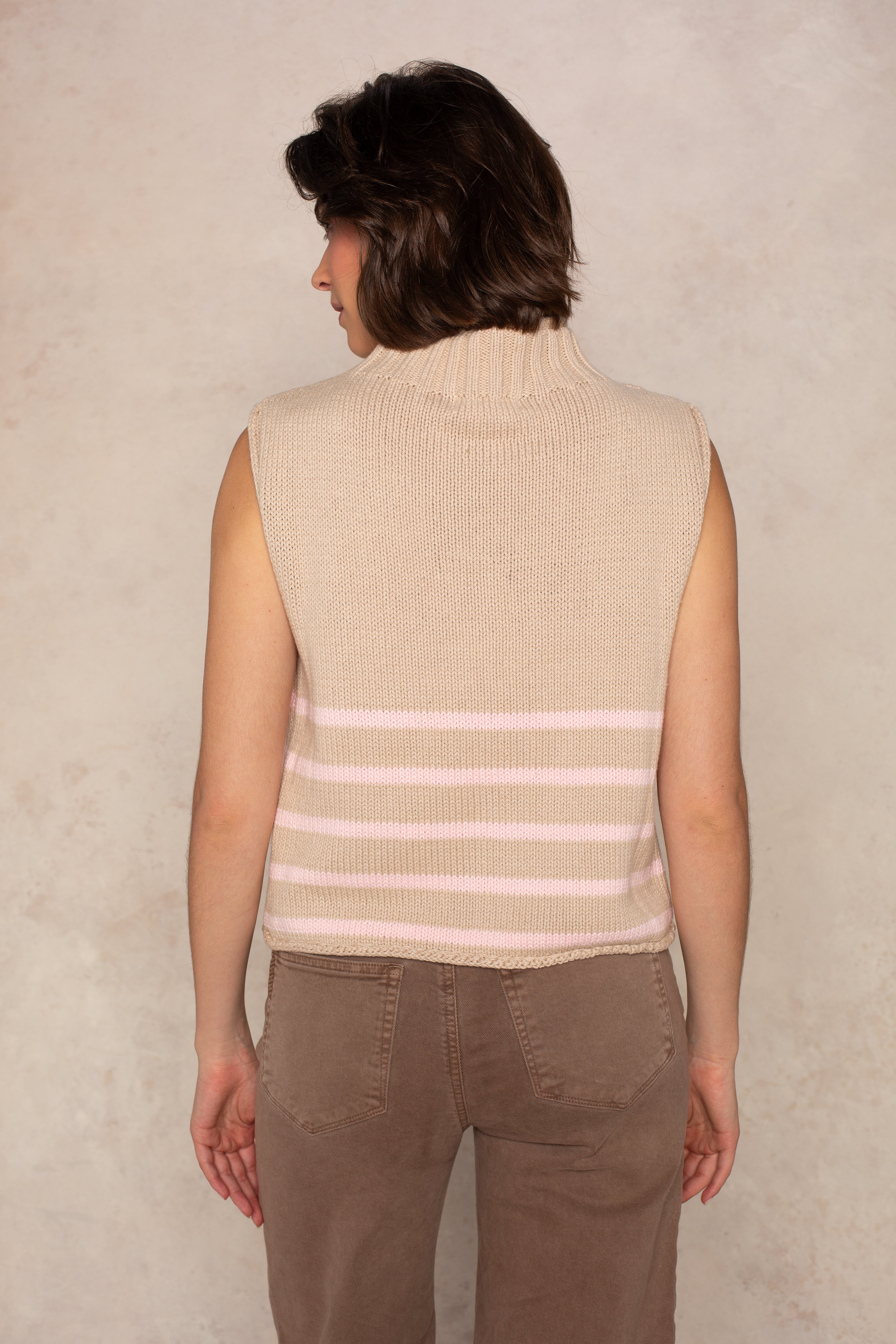 A person with short brown hair is seen from behind wearing the Georgie Grace Rolled Neck Sleeveless Sweater in Tan/Pink, paired with brown pants, against a light textured background.