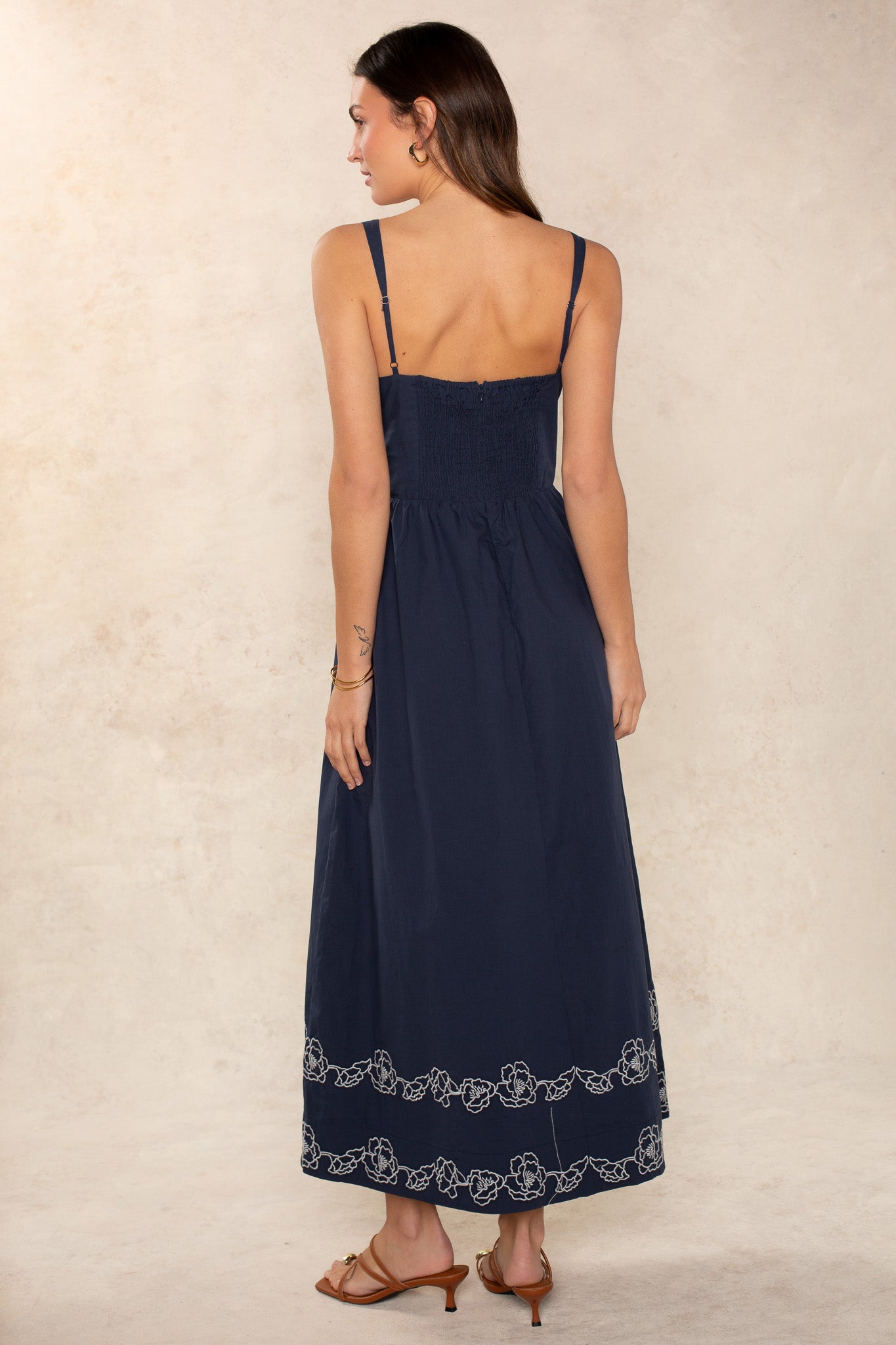A woman with long brown hair faces away, wearing the Georgie Hadley Embroidered Midi Dress in navy blue, featuring white floral embroidery along the hem and shoulder straps. She pairs it with brown sandals against a light beige background.