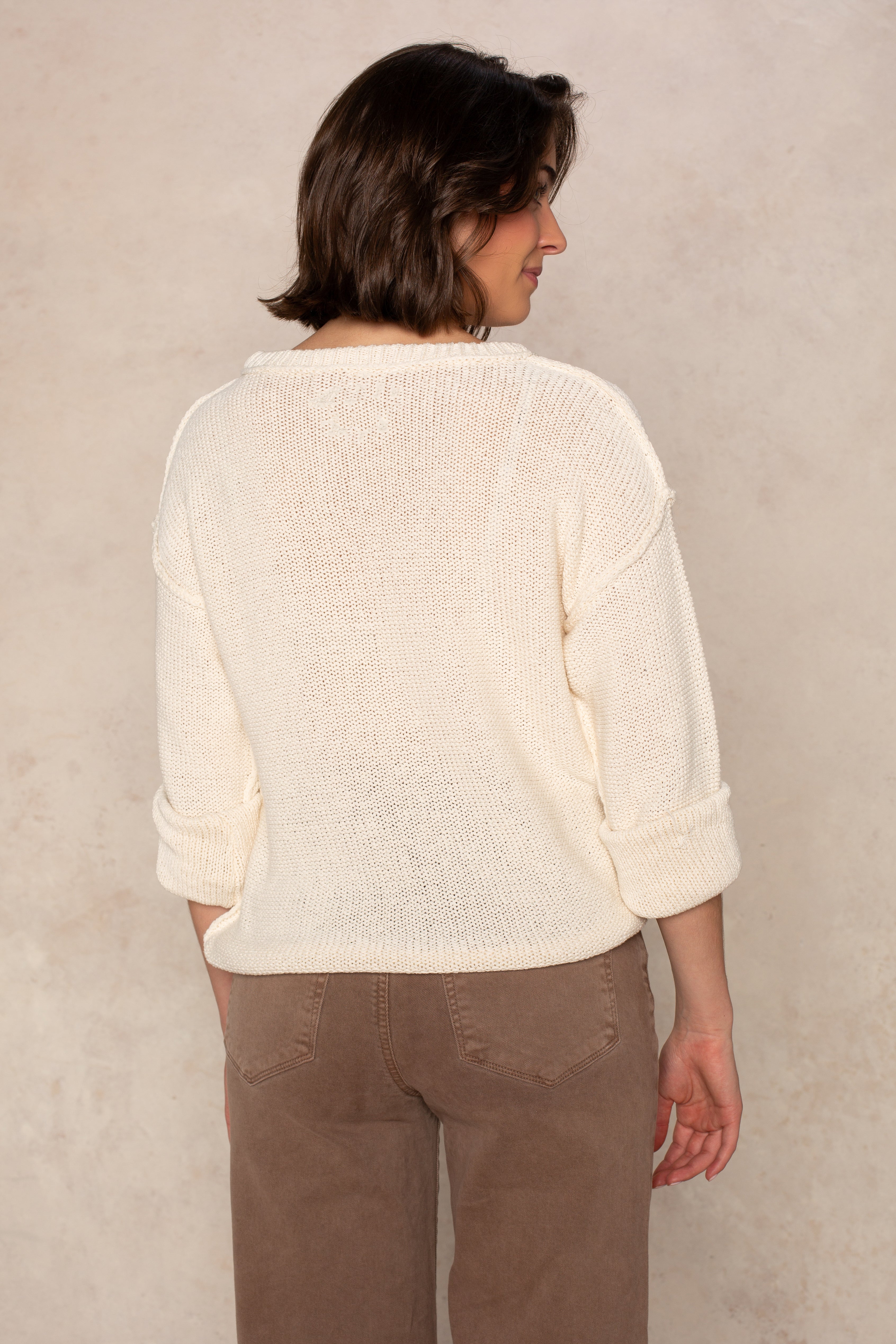 A person, facing away from the camera, wears the Isla Emmie Sweater—a soft knit with rolled-up sleeves—paired with brown pants, standing against a light beige textured background.