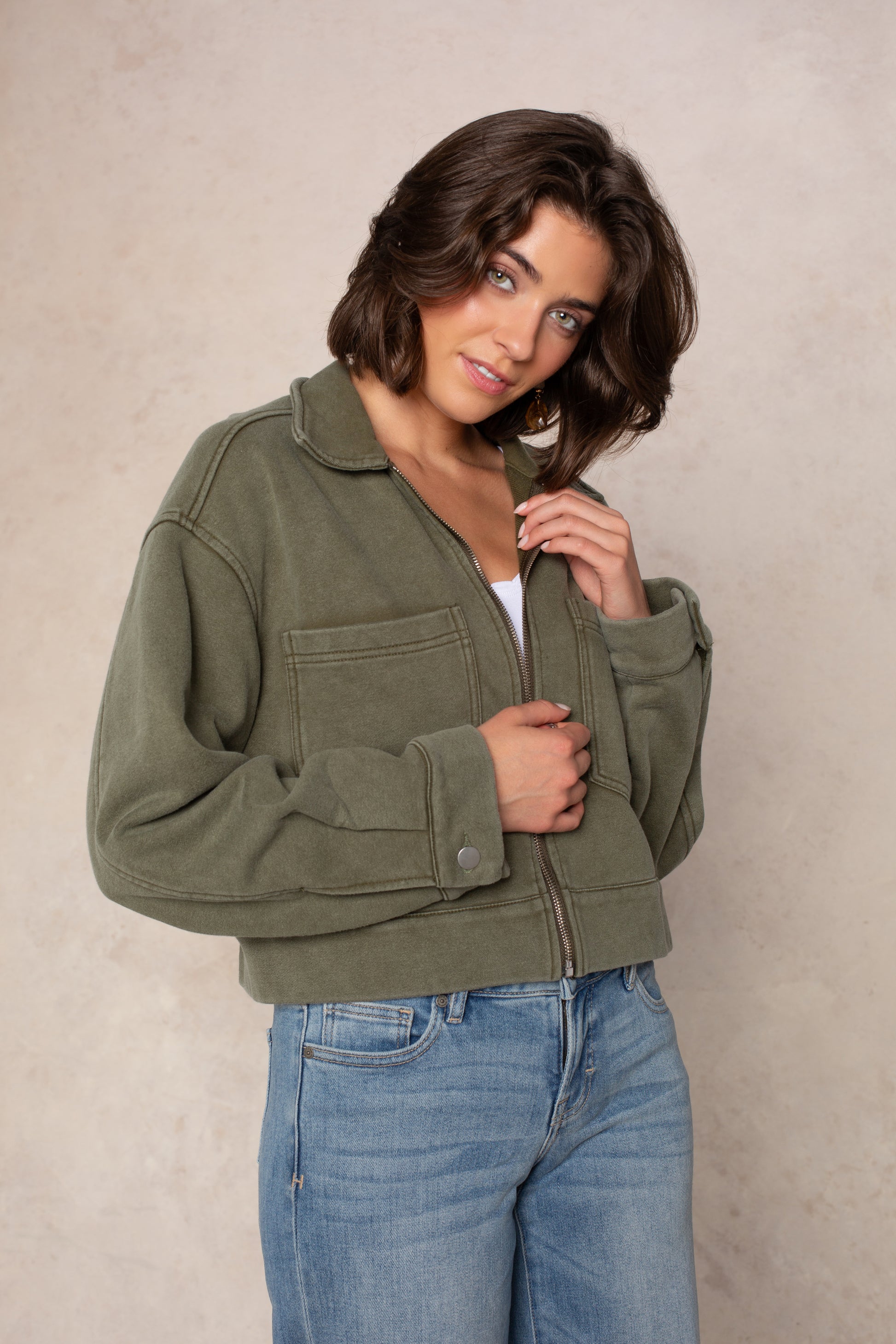 A woman with shoulder-length brown hair wears the Isla Ali Zip Up Knit Jacket over a white top and blue jeans, standing against a light beige background. She looks at the camera with a slight smile and holds the collar of her jacket.