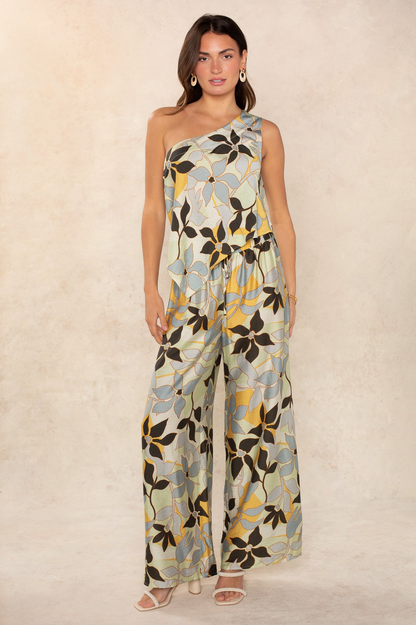 A woman stands facing forward in a modern style, wearing The Nora Set Pant by Georgie—an asymmetrical one-shoulder jumpsuit with versatile pants and a colorful abstract floral print in yellow, blue, and brown. She has long hair, sandals, and earrings.