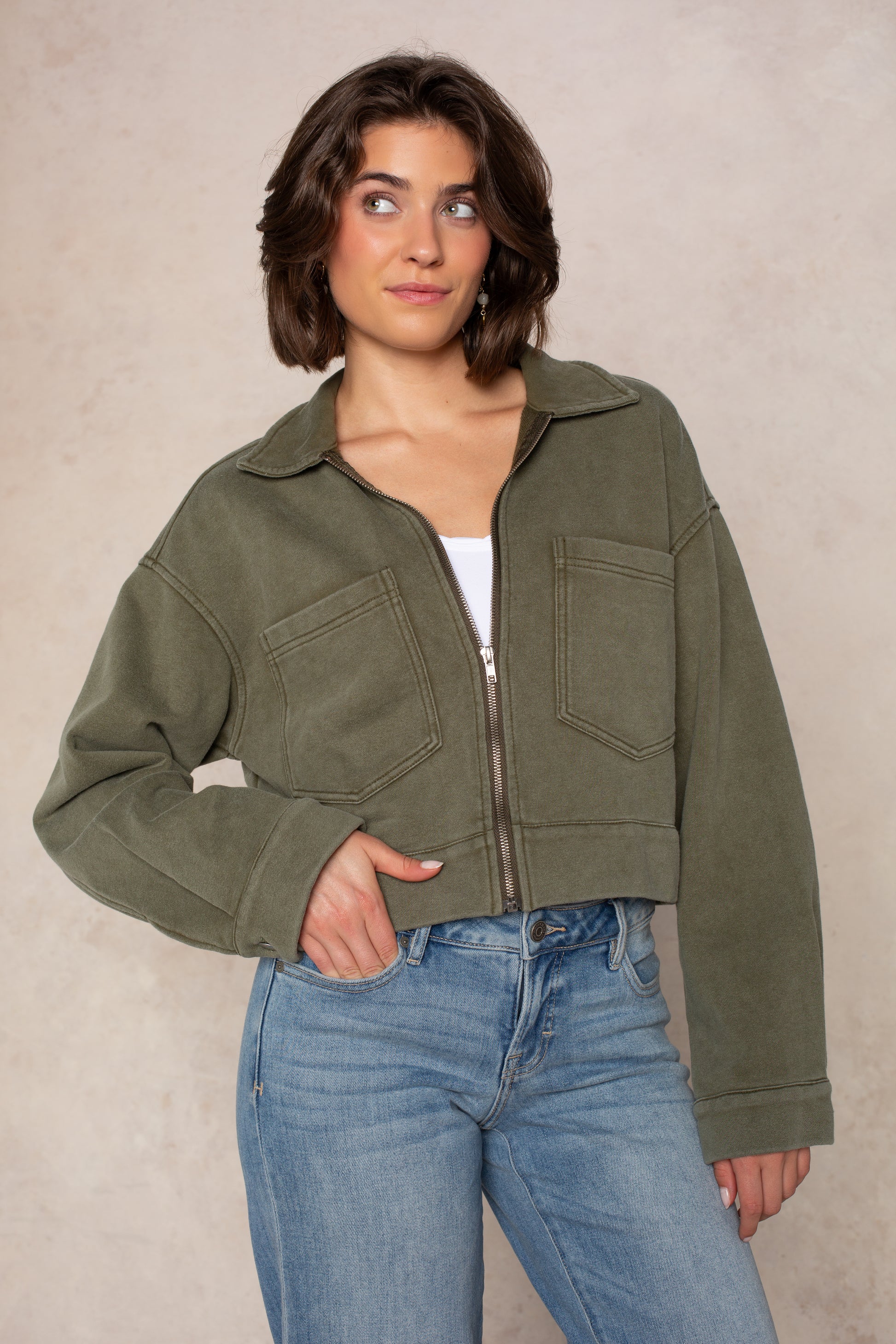 A woman with short brown hair wears the Isla Ali Zip Up Knit Jacket in Washed Olive over a white top and blue jeans, standing against a light beige background and looking up slightly with a gentle smile.