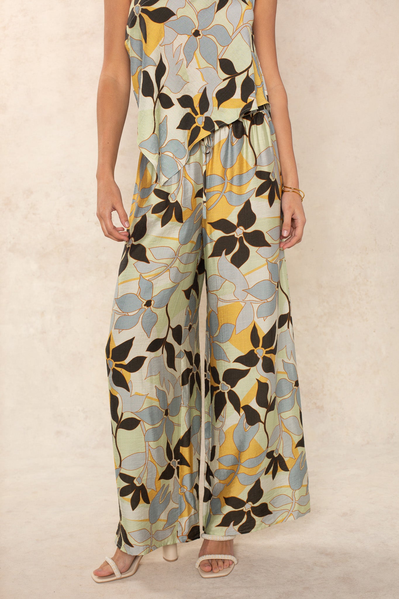 Wearing Georgie's The Nora Set Pant—wide-leg, high-waisted pants—and a matching sleeveless top with a bold yellow, black, green, and blue abstract leaf print, the look is finished with white sandals and a gold bracelet.