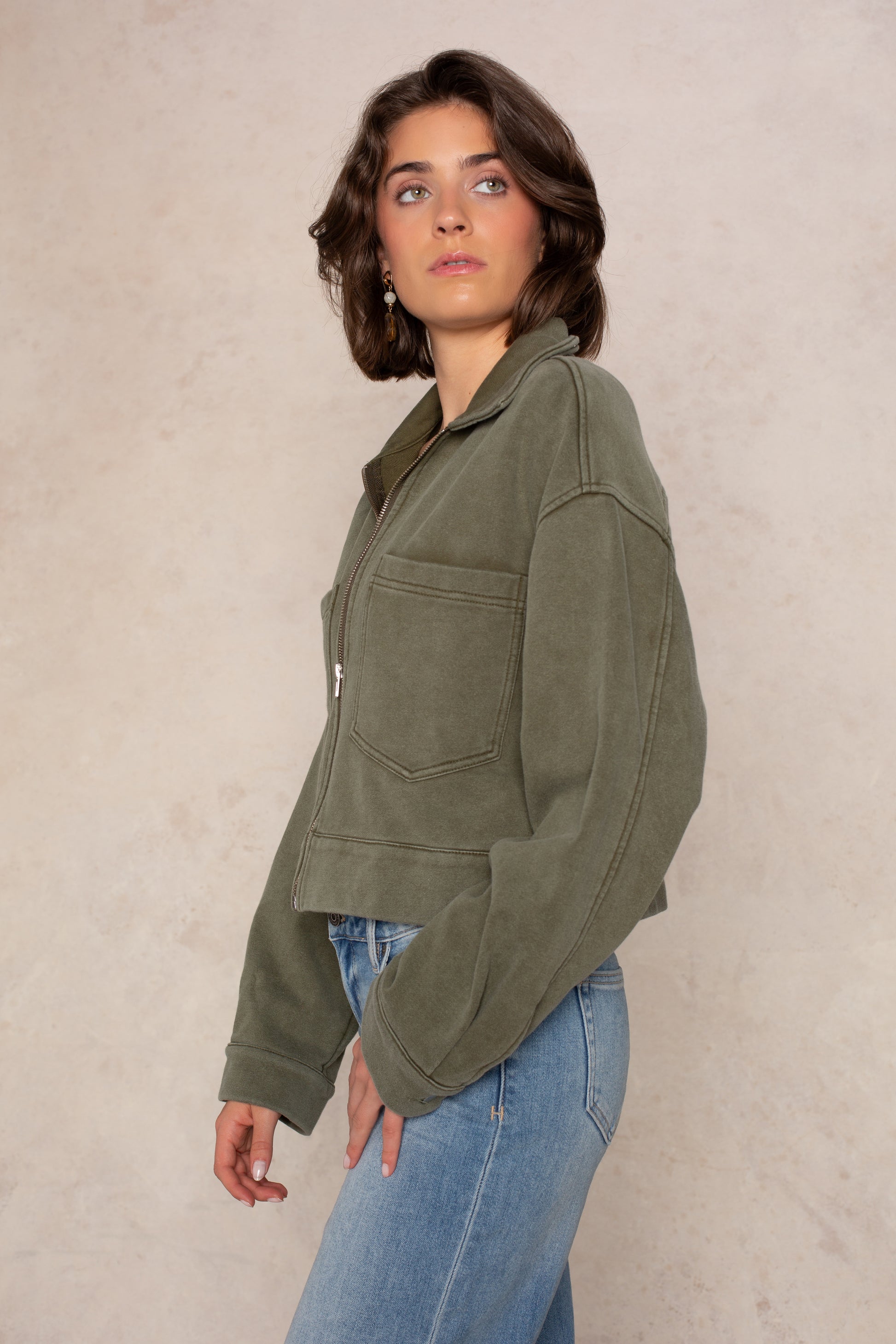 A person with short brown hair wearing the Isla Ali Zip Up Knit Jacket in olive green and blue jeans stands against a beige background, looking to the side.