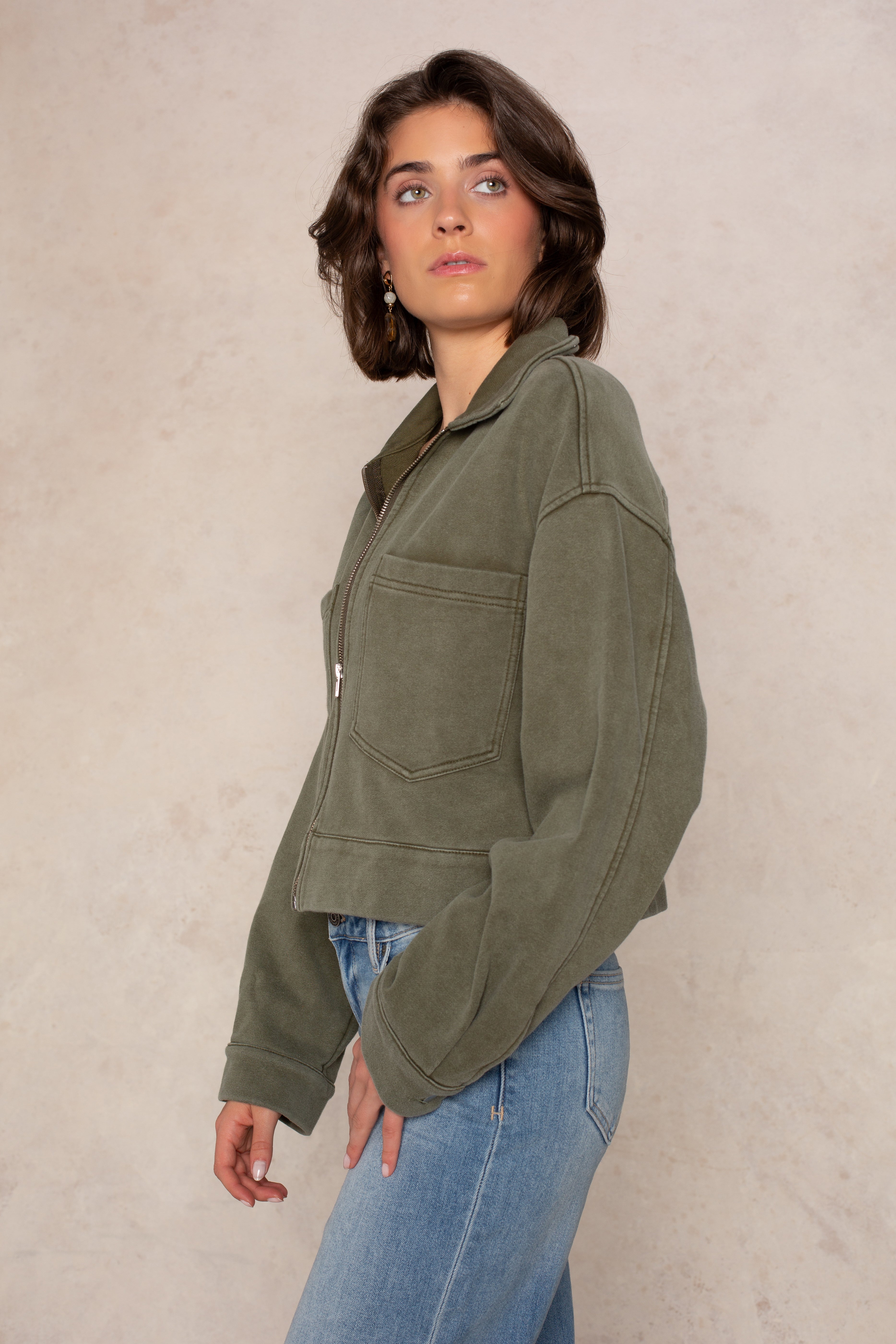 A person with short brown hair wearing the Isla Ali Zip Up Knit Jacket in olive green and blue jeans stands against a beige background, looking to the side.
