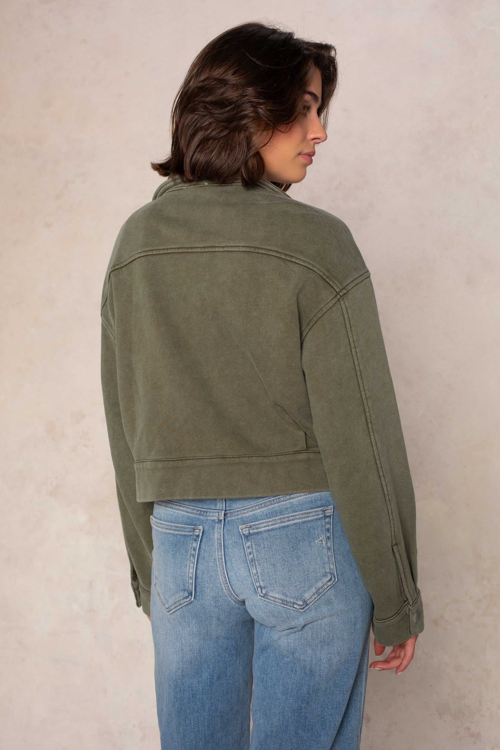 A person with short brown hair is standing, facing away. They are wearing the Isla Ali Zip Up Knit Jacket in olive green along with light blue jeans, set against a neutral beige background.