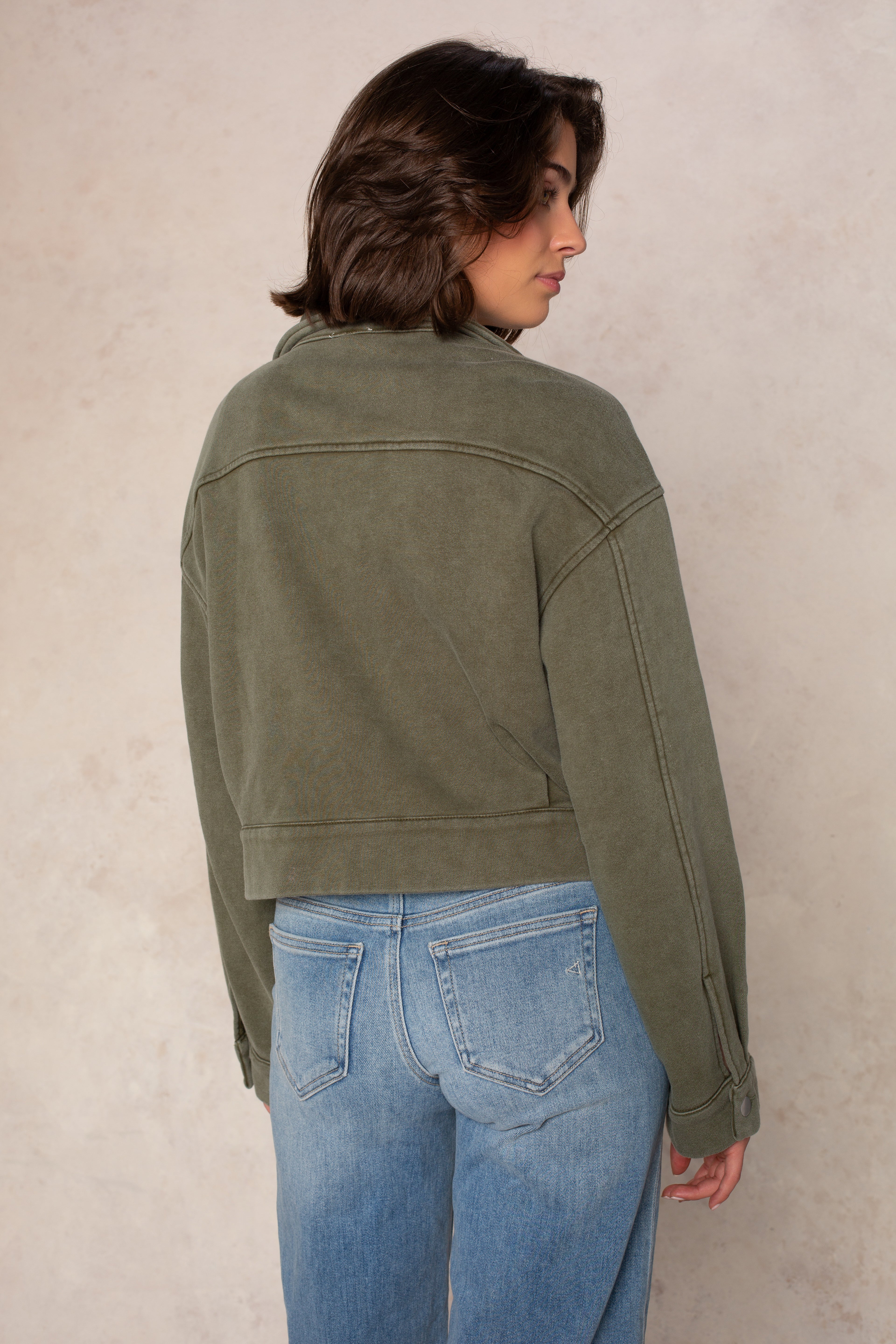 A person with short brown hair is standing, facing away. They are wearing the Isla Ali Zip Up Knit Jacket in olive green along with light blue jeans, set against a neutral beige background.
