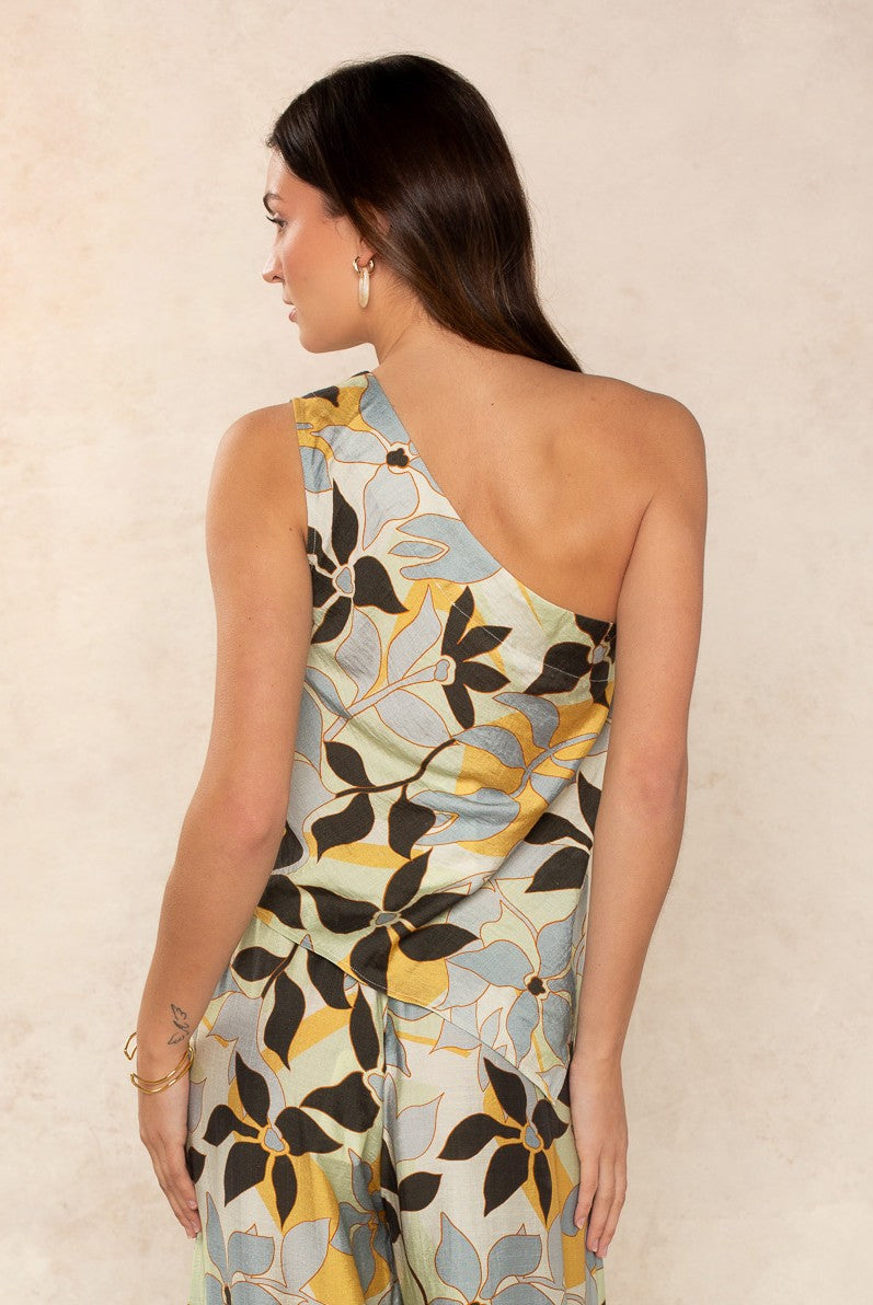 A woman with long brown hair stands sideways, wearing The Nora Set Top by Georgie. This sleeveless, one-shoulder piece features a bold abstract leaf pattern—an instant modern staple that stands out against a neutral light beige background.