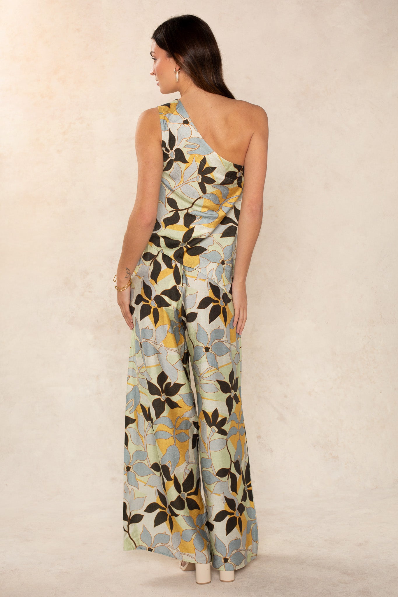 A woman faces away, wearing The Nora Set Pant by Georgie—one-shoulder, wide-leg, with bold blue, yellow, and black florals—showcasing modern style against a neutral textured background.