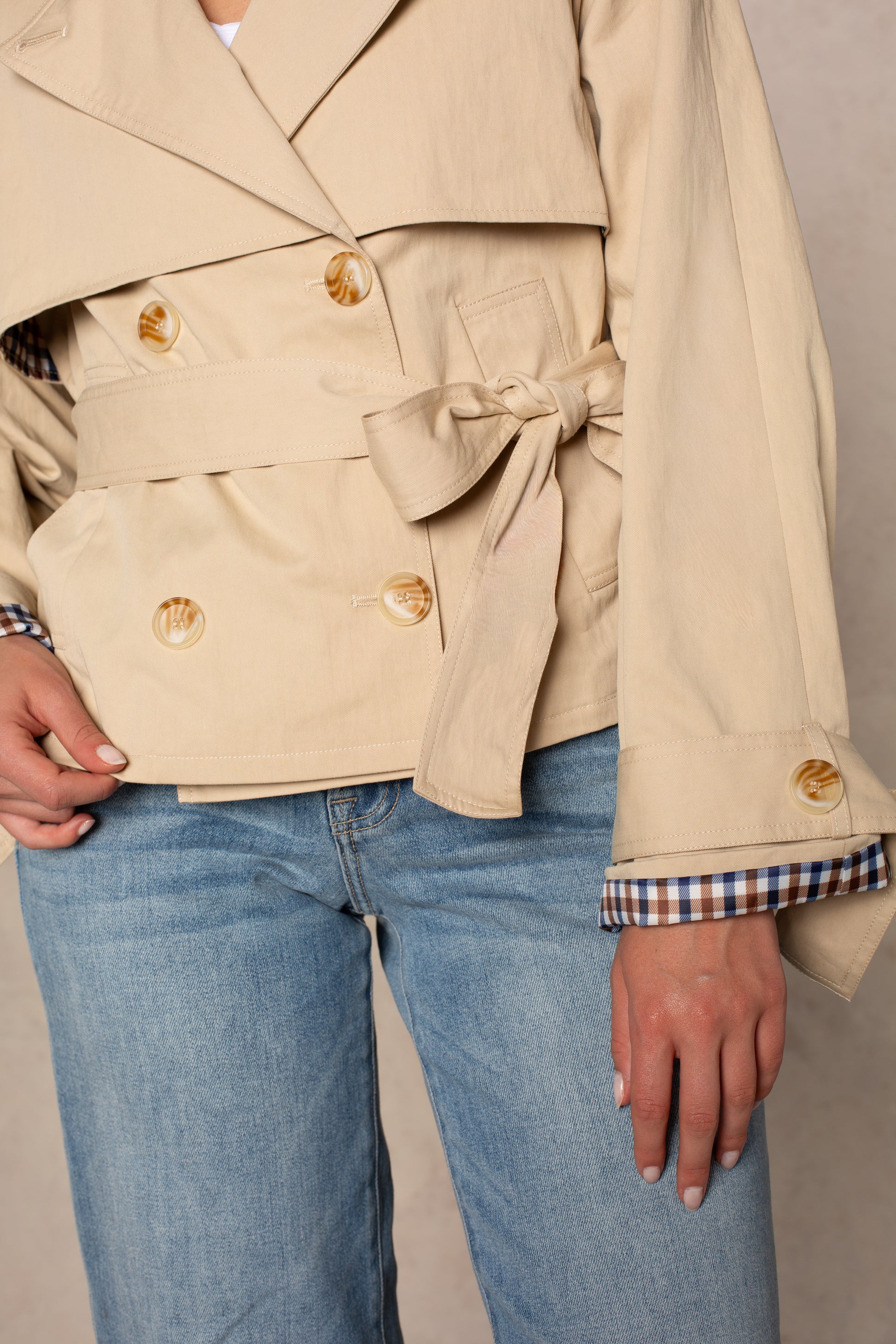 A person is seen from the torso to upper legs wearing the Georgie Carolyn Cropped Trench—a double-breasted beige coat with large buttons, belted waist, and plaid-lined cuffs—paired with light blue jeans.