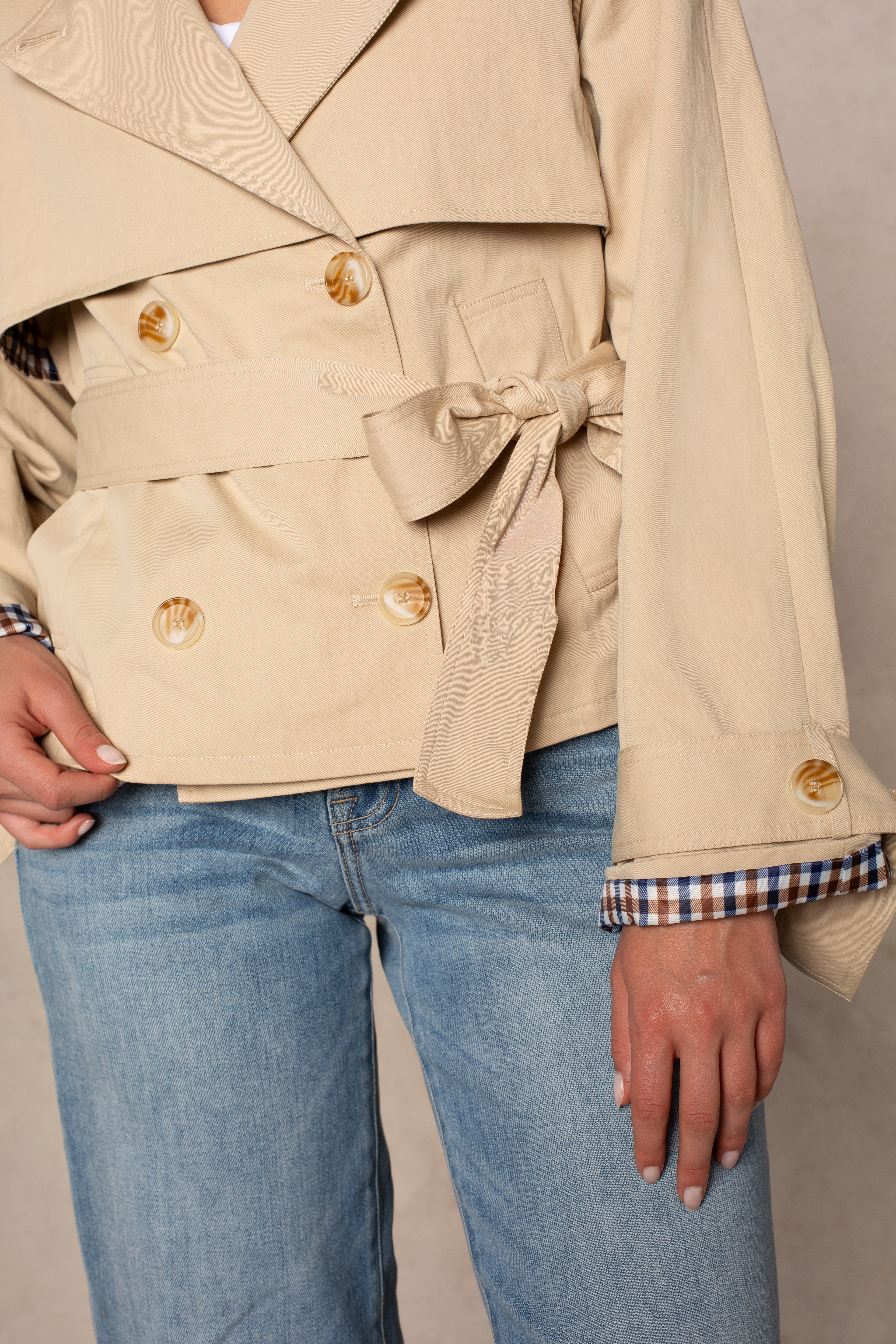 A person is seen from the torso to upper legs wearing the Georgie Carolyn Cropped Trench—a double-breasted beige coat with large buttons, belted waist, and plaid-lined cuffs—paired with light blue jeans.