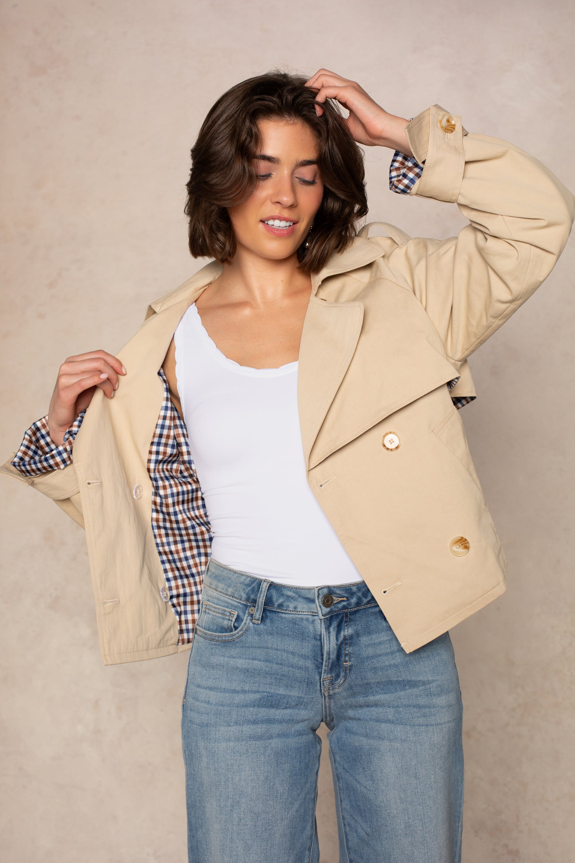A woman poses with one hand in her hair and the other holding open her Carolyn Cropped Trench by Georgie, revealing its plaid lining over a white top and blue jeans, set against a light, neutral background.