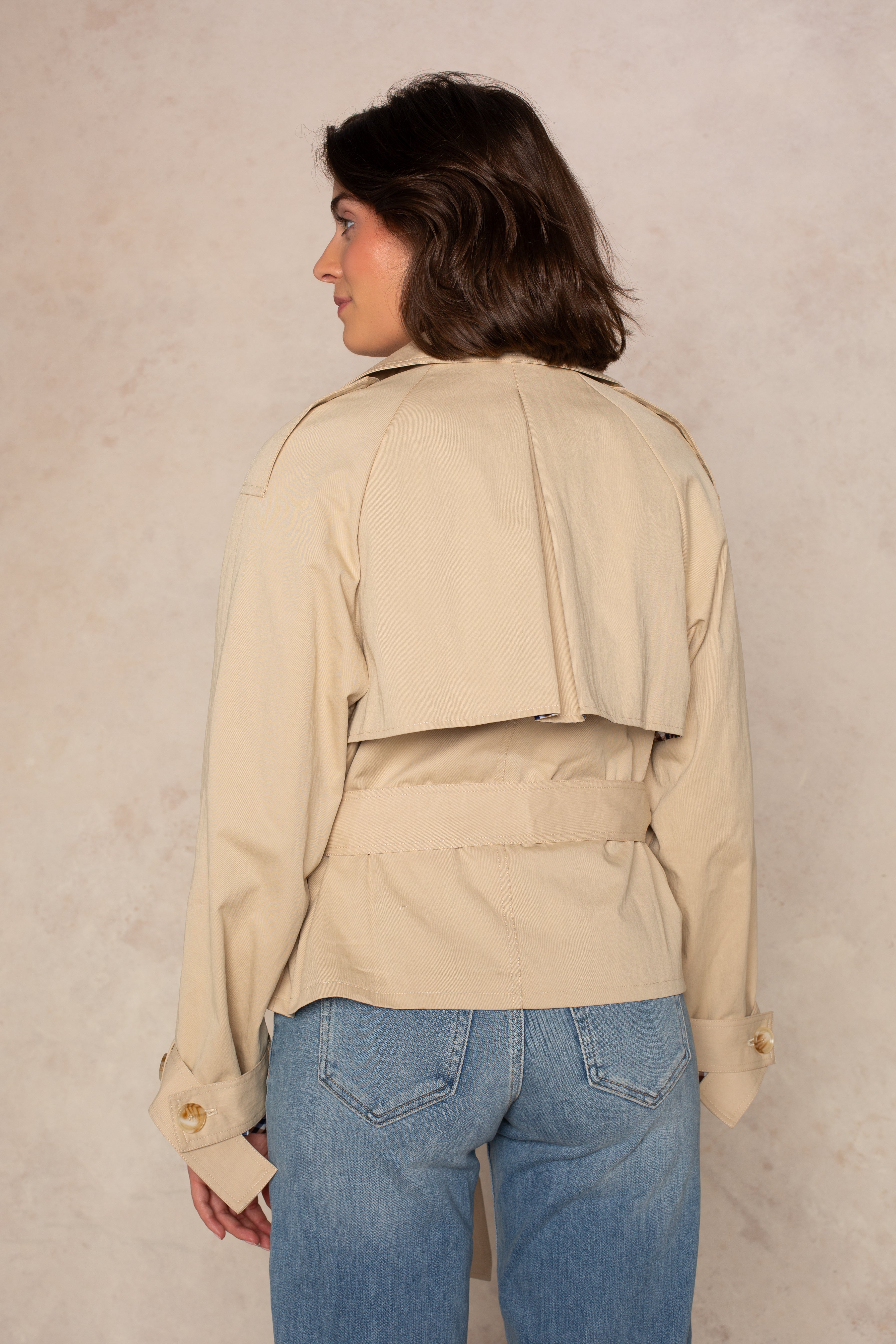 A woman with short brown hair is seen from behind, wearing Georgie's Carolyn Cropped Trench and blue jeans, standing against a light, textured background.