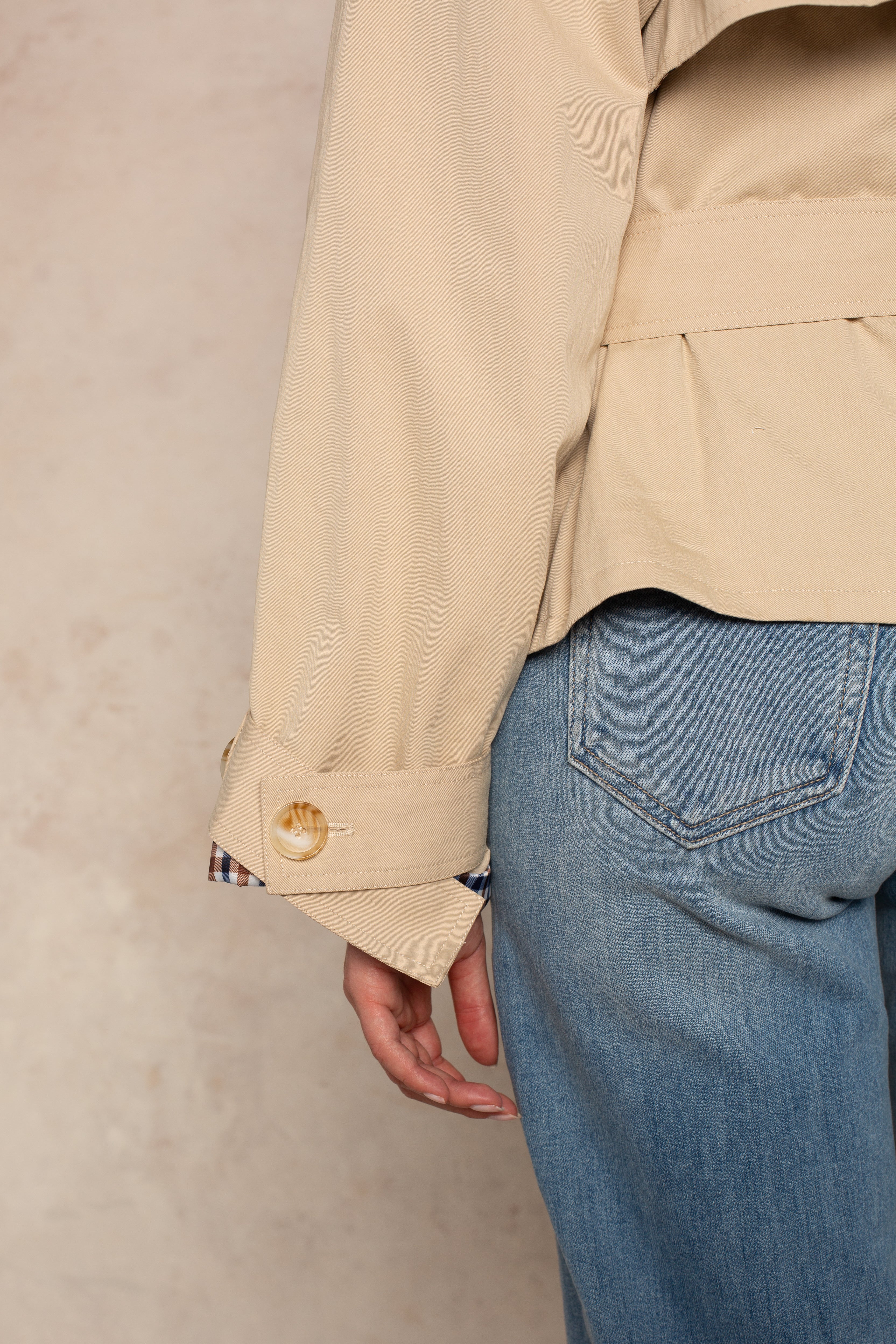 Only the lower back, arm, and hand are visible as a person models the Georgie Carolyn Cropped Trench with large buttoned cuffs, paired with light blue jeans against a neutral textured background.