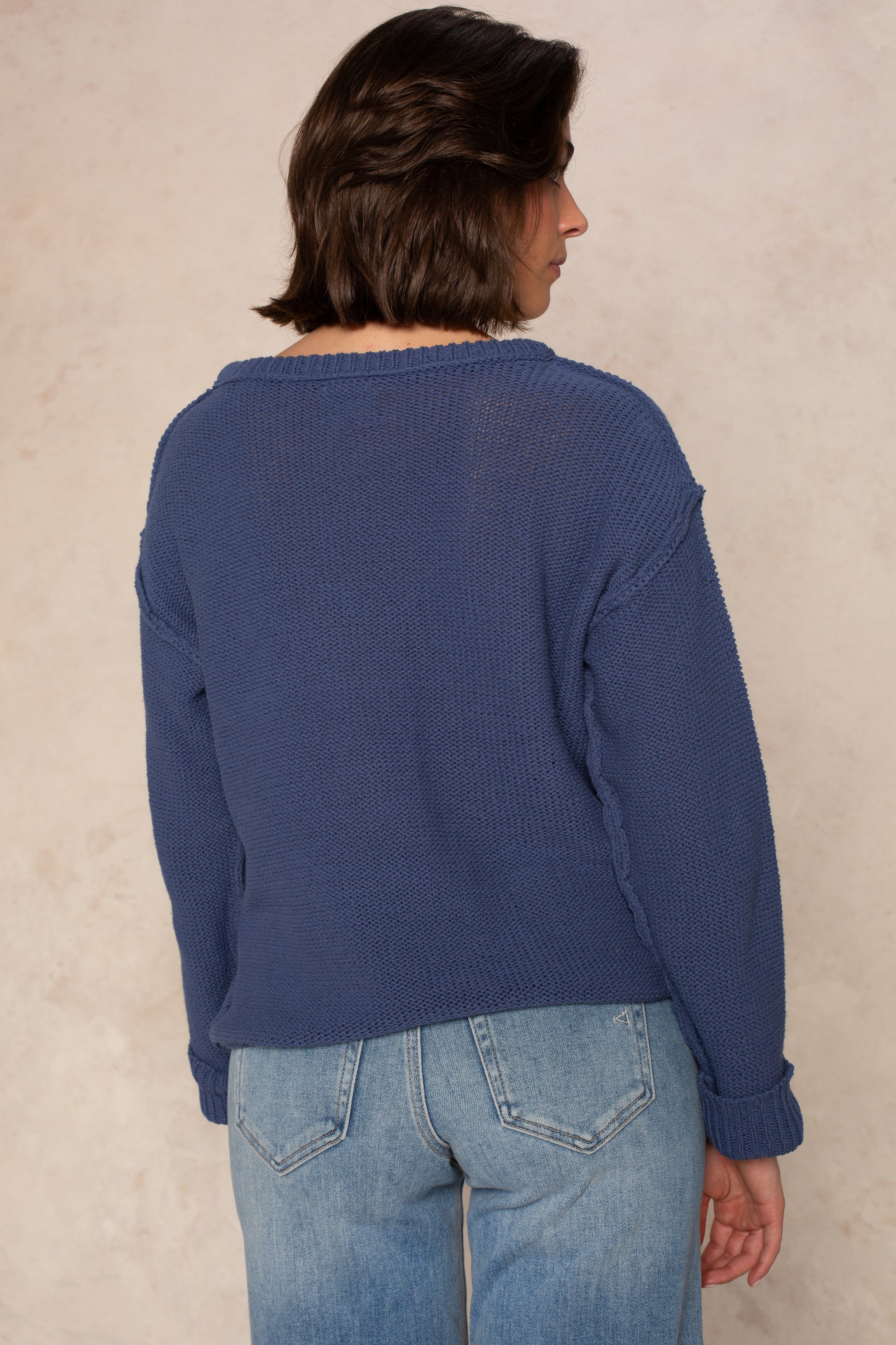 A person with shoulder-length brown hair stands with their back to the camera against a beige background, wearing Isla's Emmie Sweater in light blue paired with light blue jeans.
