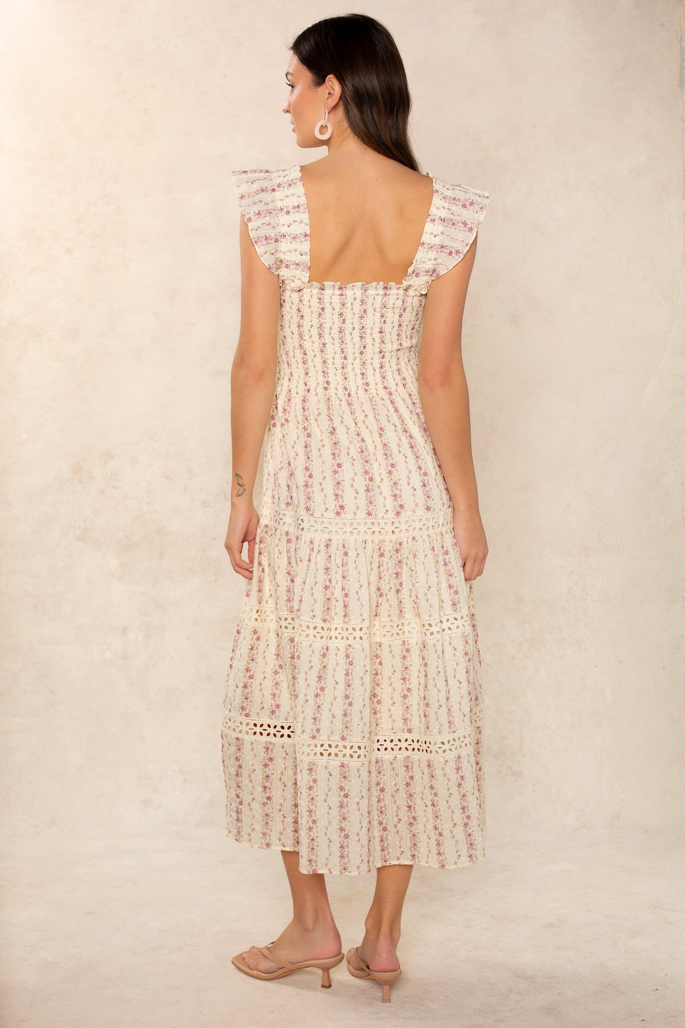 A woman with long brown hair stands facing away, wearing the Georgie Calypso Smocked Eyelet Midi Dress in cream with pink floral patterns, ruffled sleeves, and lace accents. She pairs it with nude heels and white circular earrings.