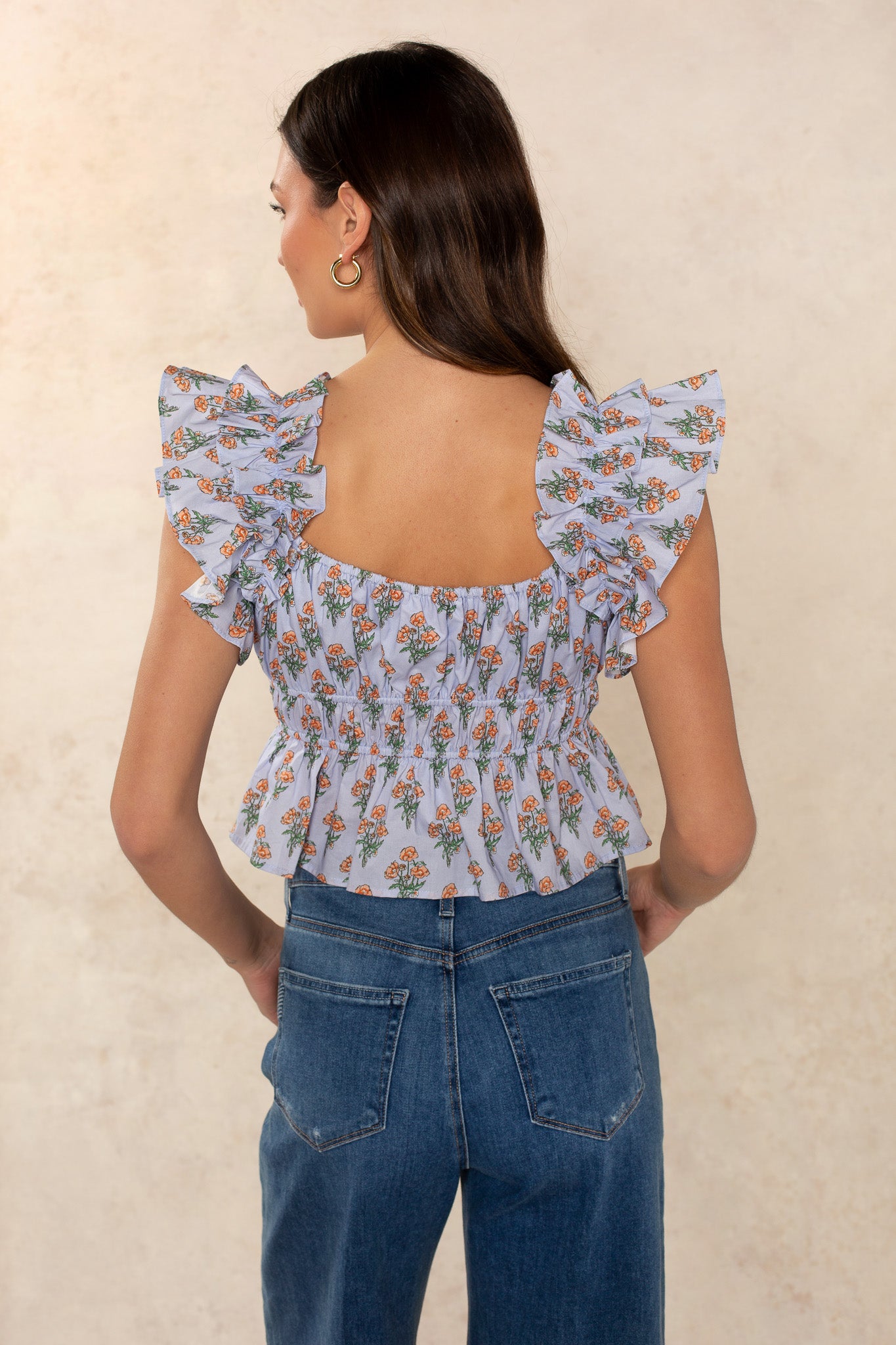 A woman with long brown hair wears the Avery Top by Georgie, a light blue floral ruffled blouse with flutter sleeves and a refined silhouette, paired with high-waisted blue jeans. She poses back to the camera against a beige background.