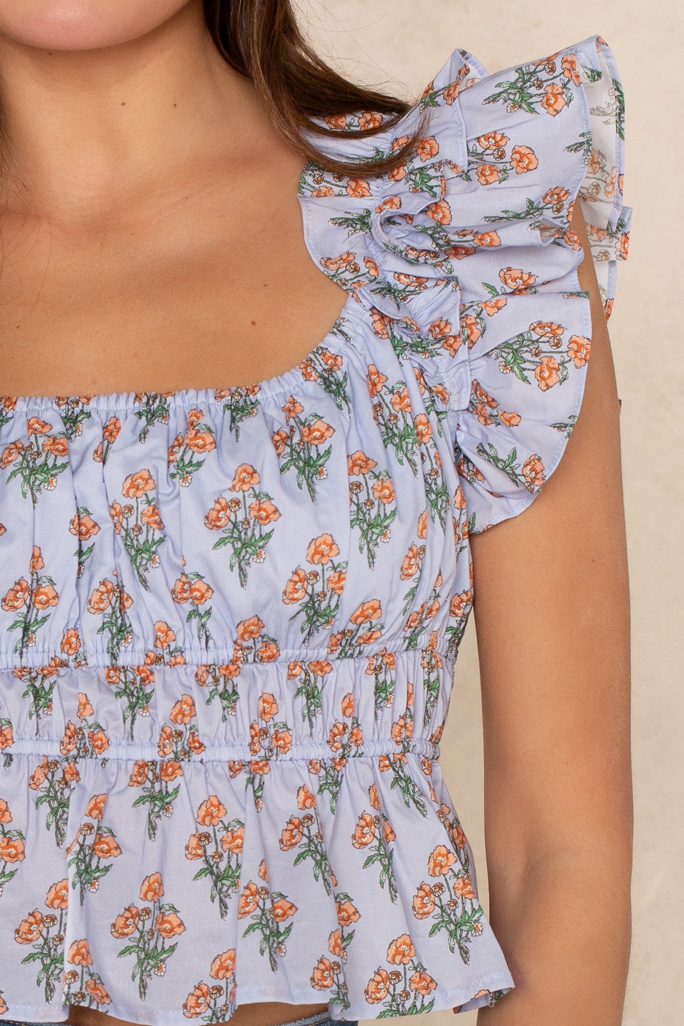 A close-up of a woman wearing the Georgie Avery Top, featuring orange flowers, ruffle sleeves, and a square neck in light purple. The image highlights her shoulders and upper chest.