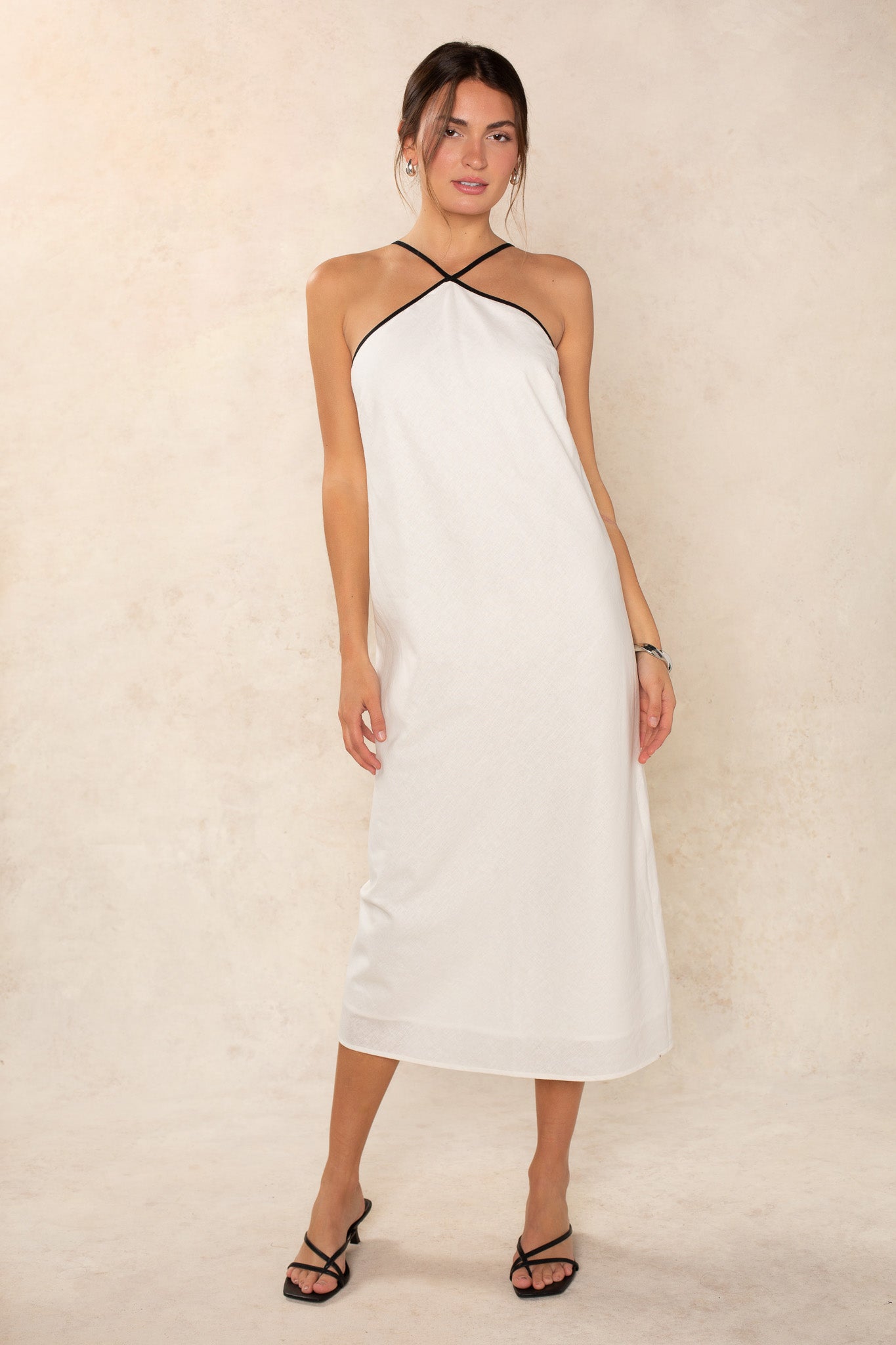 A woman poses against a beige background wearing the Georgie Margot Linen Midi Dress, featuring a sleeveless halter neckline with black trim, paired with black strappy sandals. Her hair is tied back and one hand rests at her side.