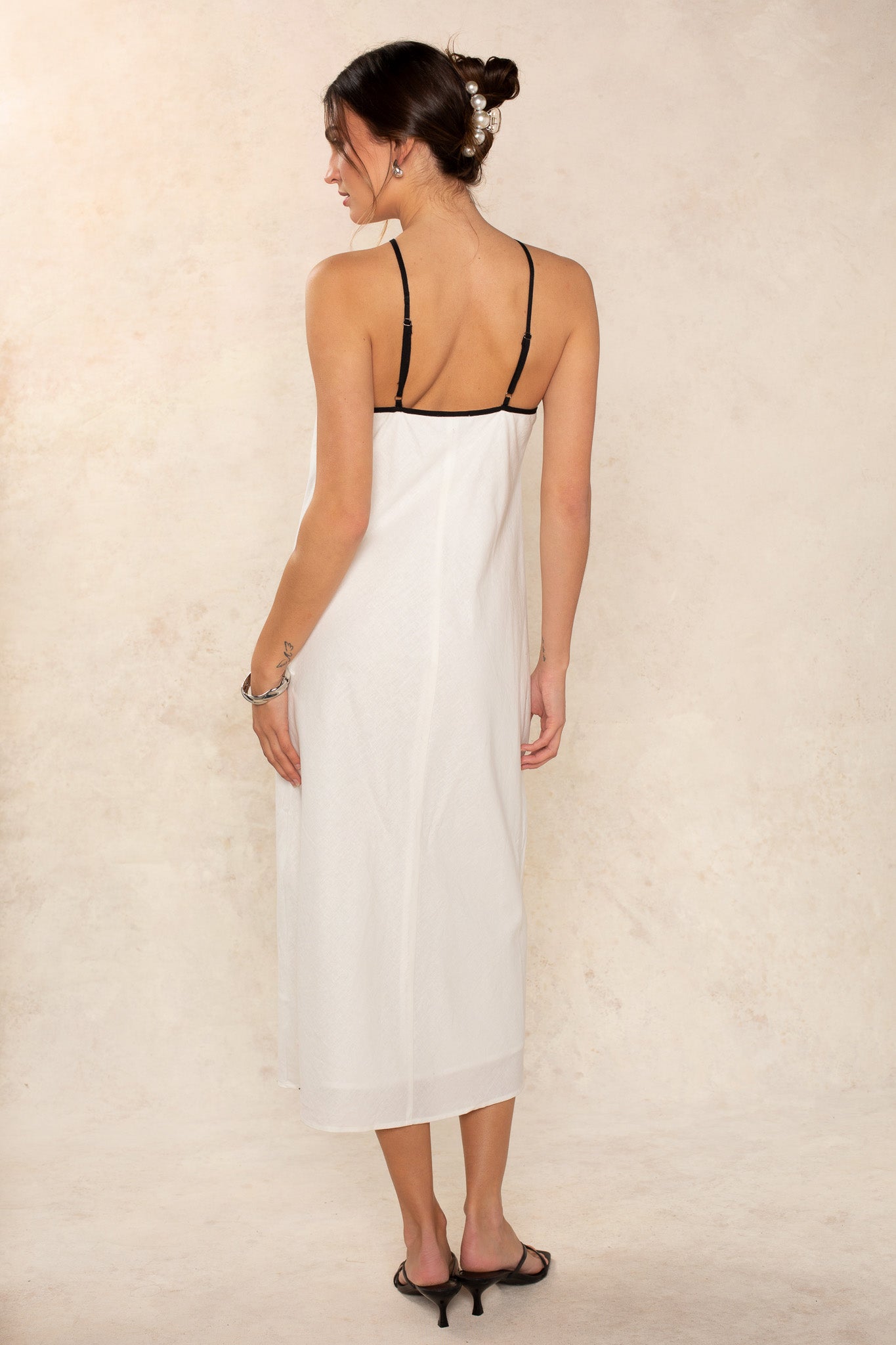 A woman faces away from the camera in a Georgie Margot Linen Midi Dress with thin black straps and black heeled sandals. Her hair is in a bun with clips, set against a neutral, textured background.