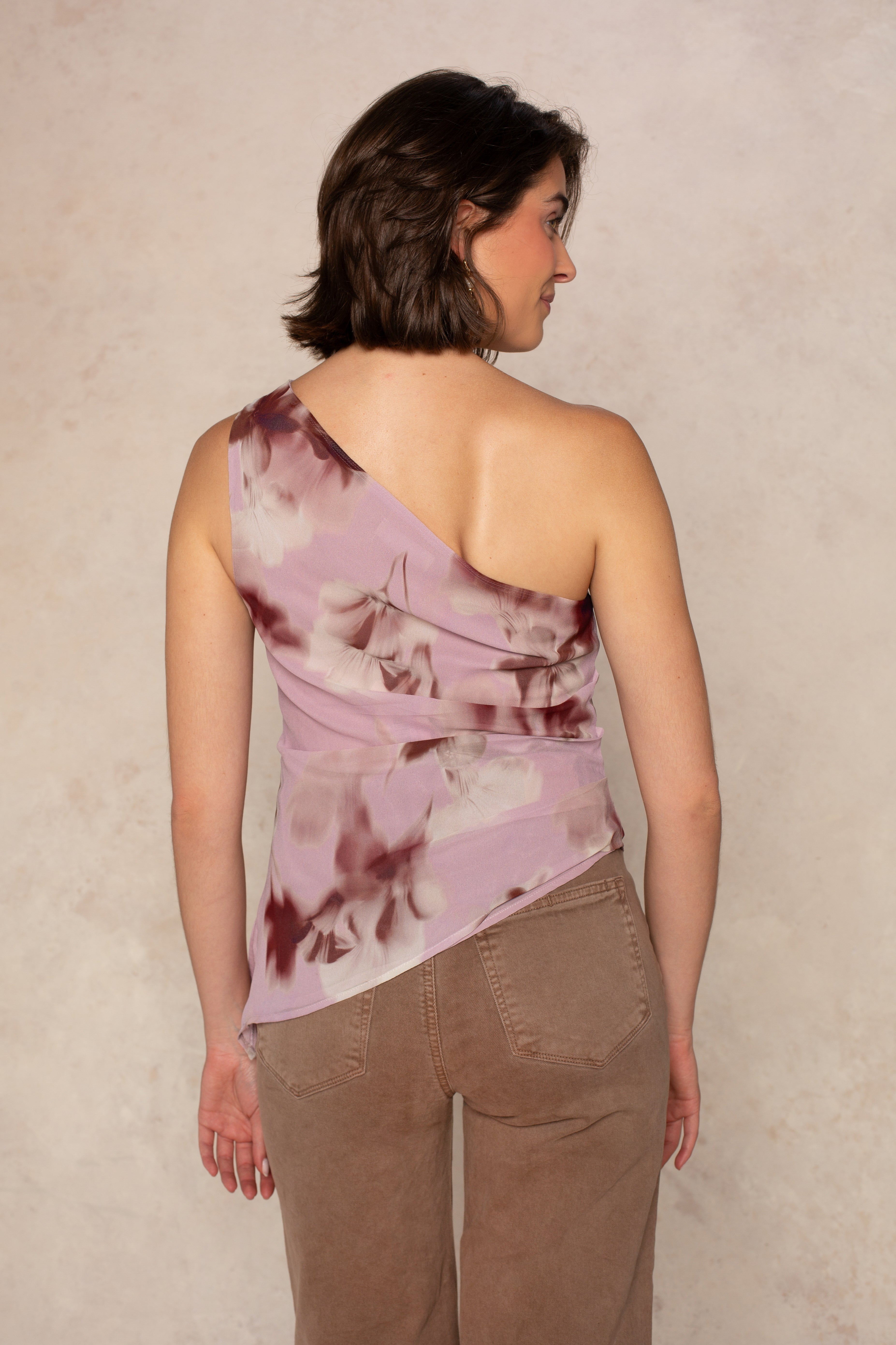 A woman with short brown hair faces away, wearing the Georgie Annabelle One Shoulder Satin Top with a floral print and asymmetrical hem, paired with light brown pants, set against a neutral textured background.